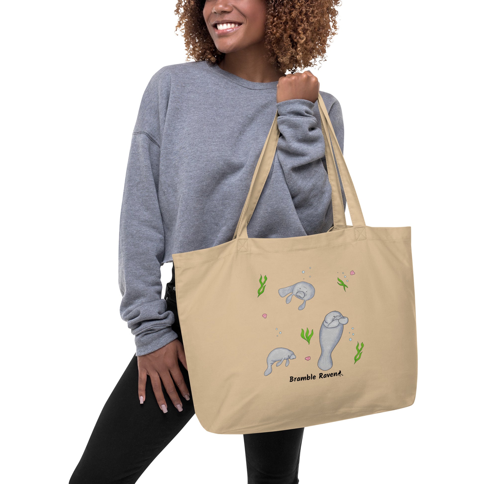 20″ × 14″ × 5″ 100% organic cotton tote bag. Features three manatees with accents of seashells, seaweed, and bubbles. Oyster colored fabric with dual handles. Shown hanging from female model's hand.
