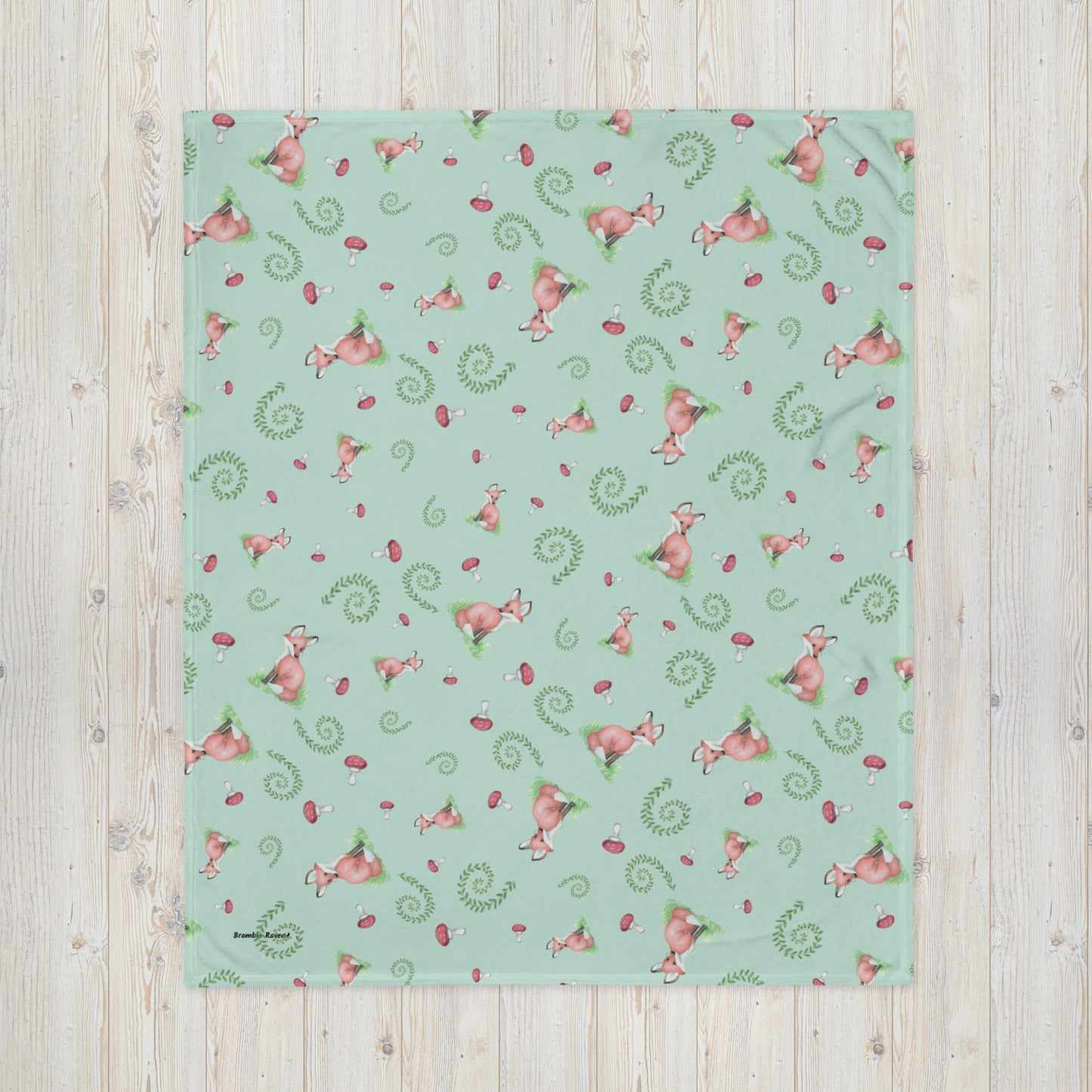 50 by 60 inch watercolor forest fox patterned blanket. Made of soft and fluffy polyester. Has foxes, mushrooms, and ferns on light green background. White underside. Machine-washable, hypoallergenic, and flame retardant. Shown on floor.