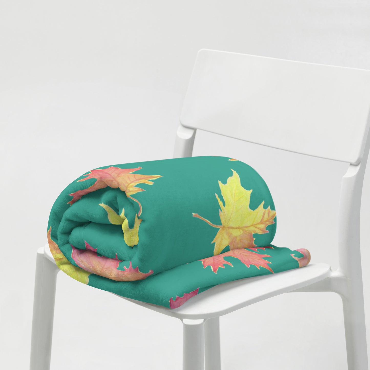50 by 60 inch soft throw blanket featuring watercolor fall leaves patterned on a dark green background. Shown rolled up on a white chair.