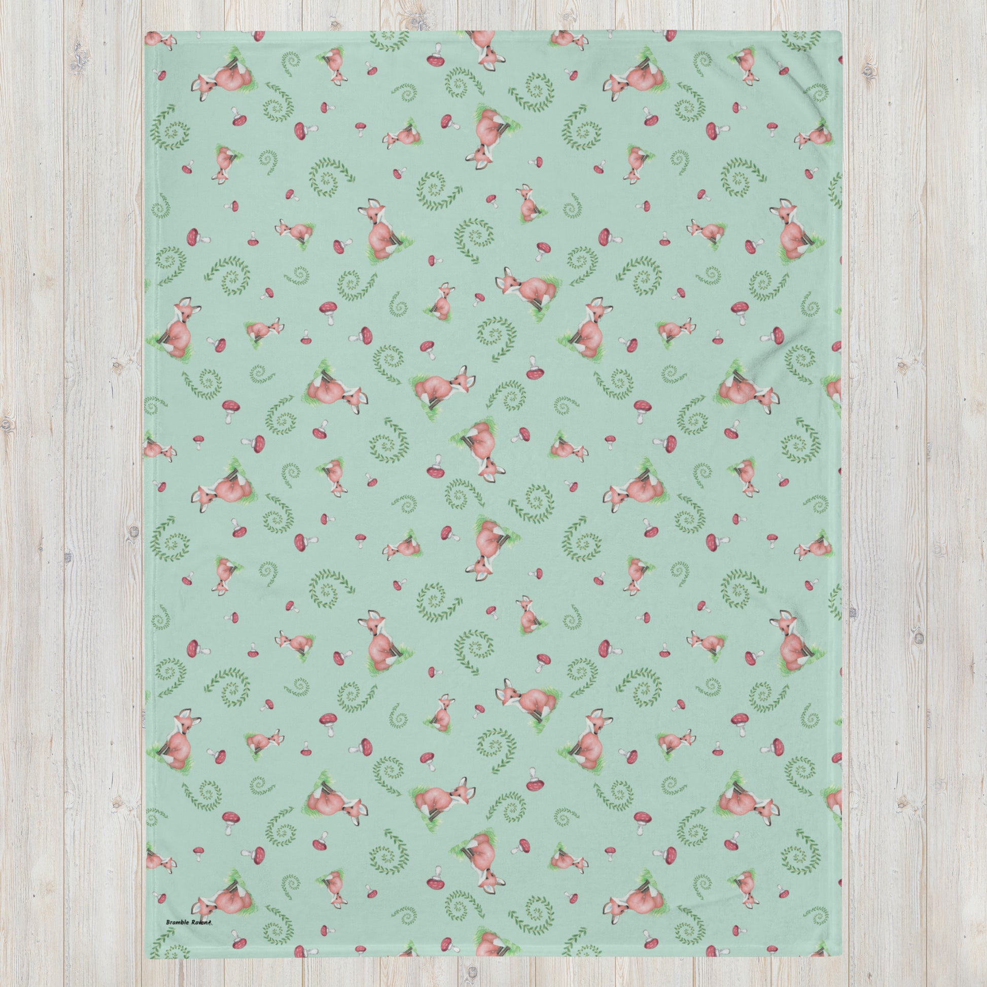 60 by 80 inch watercolor forest fox patterned blanket. Made of soft and fluffy polyester. Has foxes, mushrooms, and ferns on light green background. White underside. Machine-washable, hypoallergenic, and flame retardant. Shown on floor.