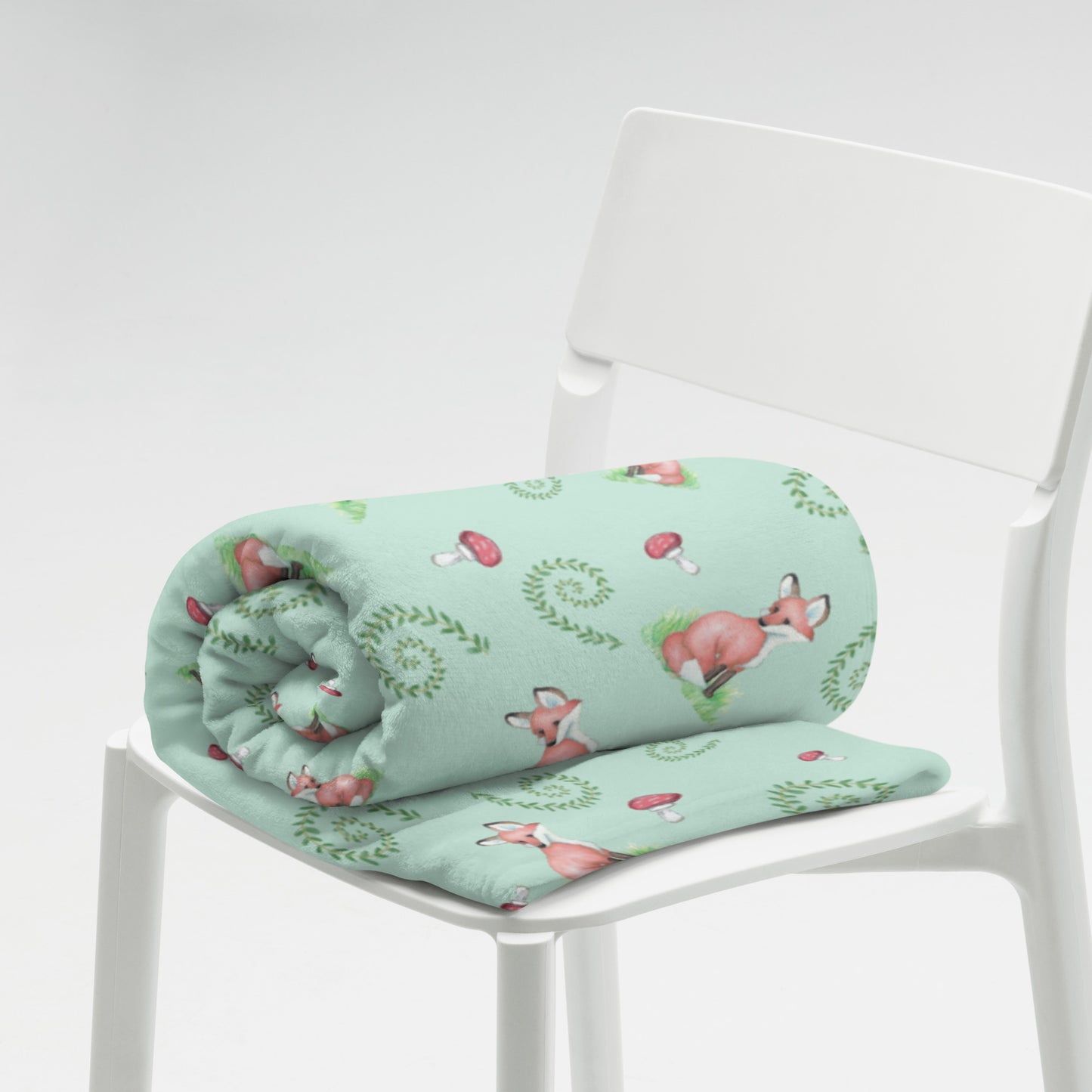 60 by 80 inch watercolor forest fox patterned blanket. Made of soft and fluffy polyester. Has foxes, mushrooms, and ferns on light green background. White underside. Machine-washable, hypoallergenic, and flame retardant. Shown rolled on a white chair.