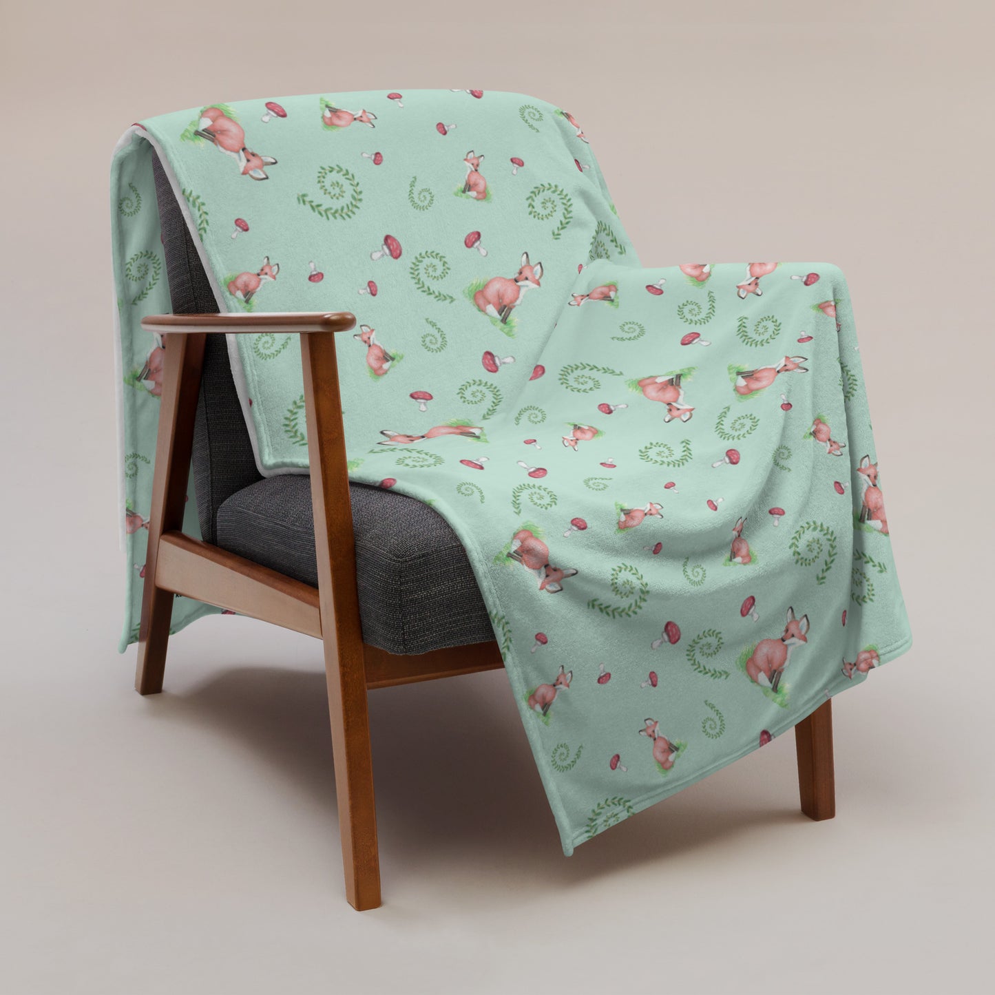 60 by 80 inch watercolor forest fox patterned blanket. Made of soft and fluffy polyester. Has foxes, mushrooms, and ferns on light green background. White underside. Machine-washable, hypoallergenic, and flame retardant. Shown on sofa.