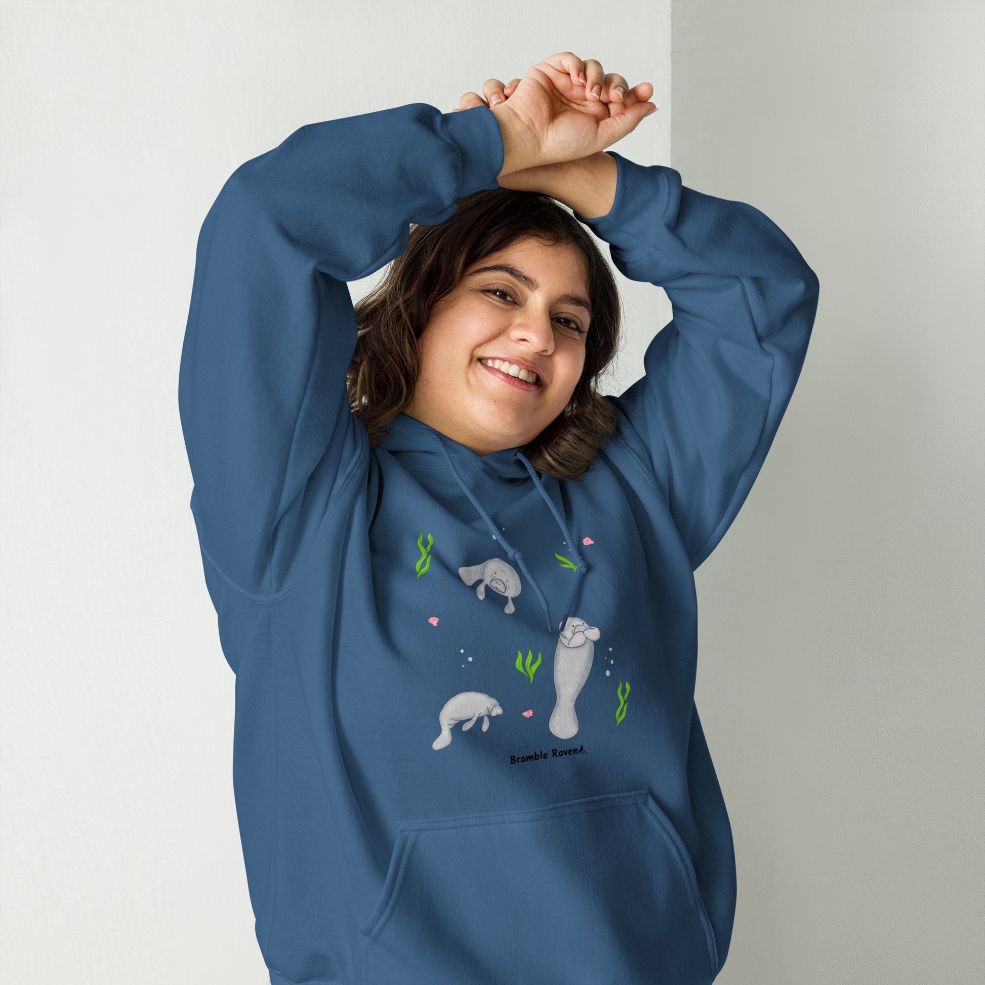 Indigo blue colored unisex hoodie. 50% cotton 50% polyester. Features a design of three manatees surrounded by seaweed, seashells, and bubbles. Has a double lined hood, ribbed cuffs, and a front pouch pocket. Shown on female model.