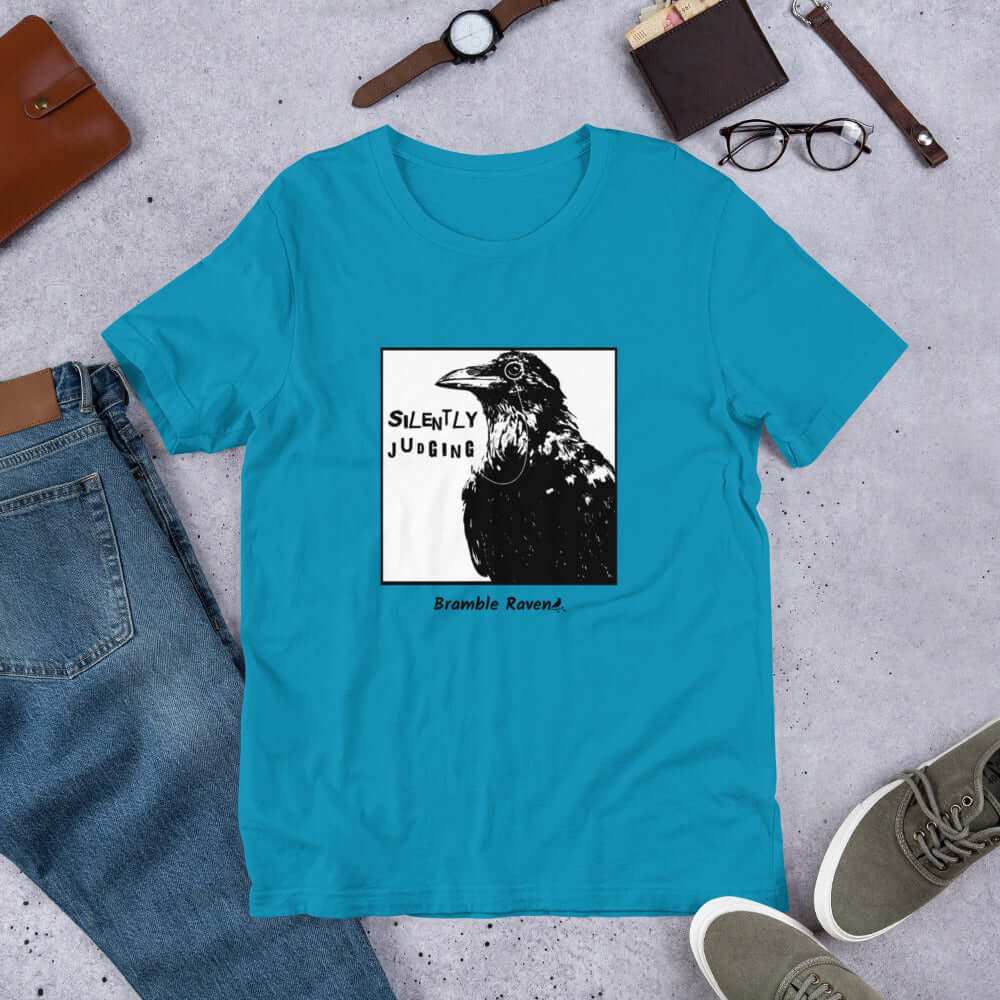 Unisex aqua colored t-shirt. Features silently judging text next to black crow wearing a monocle in a square frame on a white background. 