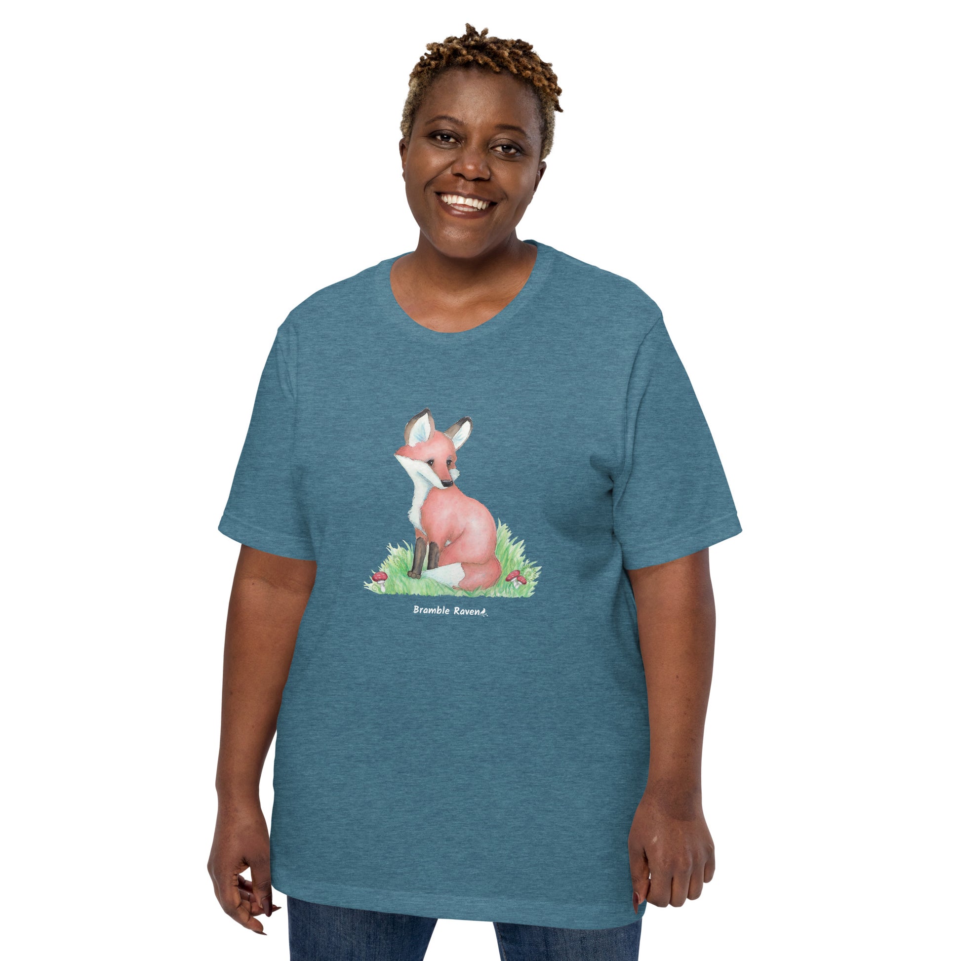 Unisex heather deep teal colored forest fox t-shirt. Features watercolor print of a fox in the grass surrounded by mushrooms and ferns. Shown on female model.