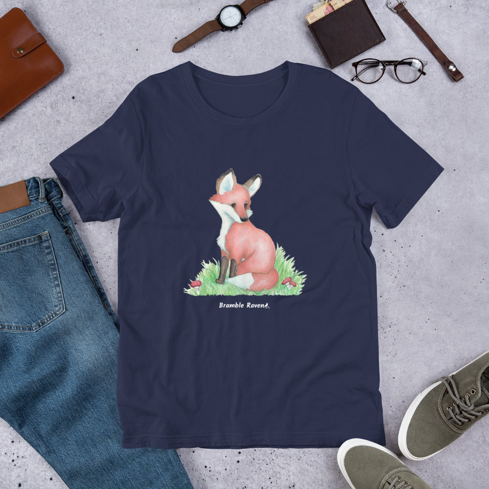 Unisex navy blue colored forest fox t-shirt. Features watercolor print of a fox in the grass surrounded by mushrooms and ferns. Shown on ground by pants, shoes, and glasses.