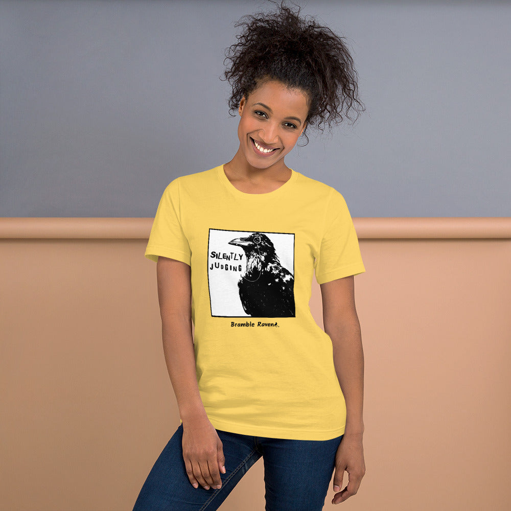 Unisex yellow colored t-shirt. Features silently judging text next to black crow wearing a monocle in a square frame on a white background.  Shown on female model.