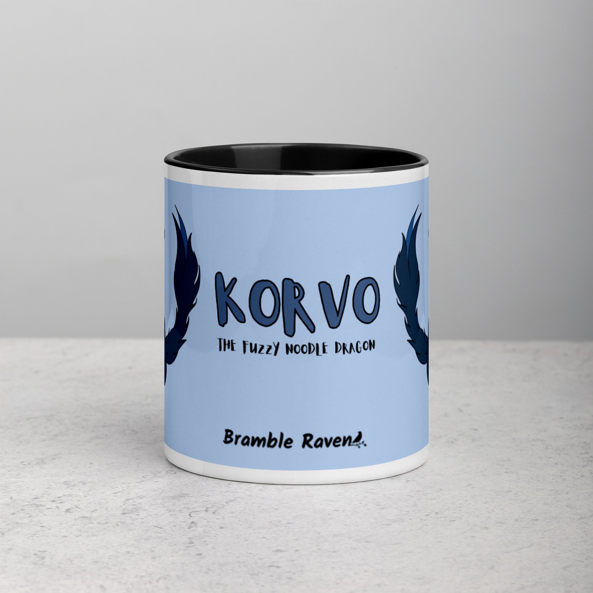 11 ounce ceramic mug featuring a double-sided image of angry Korvo the Fuzzy Noodle dragon against a light blue background.  Black inside and handle. Sitting on tabletop. Image shows front of mug with Korvo the Fuzzy Noodle Dragon text and Bramble Raven logo.
