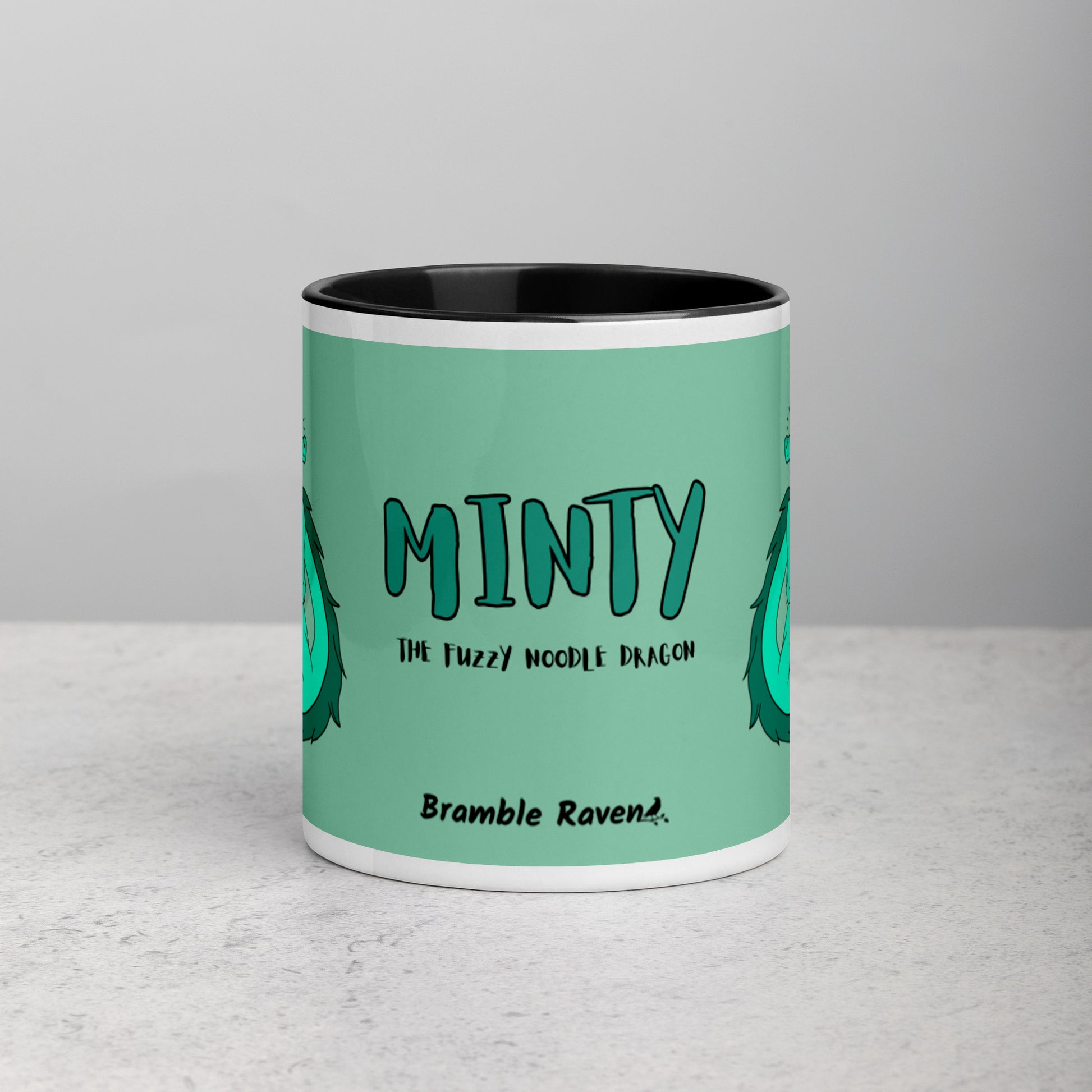 11 ounce white ceramic mug. Black inside and handle. Features double-sided image of Minty the Fuzzy Noodle Dragon on a green background. Shown on tabletop. Front view shows Minty the Fuzzy Noodle Dragon text and Bramble Raven logo.