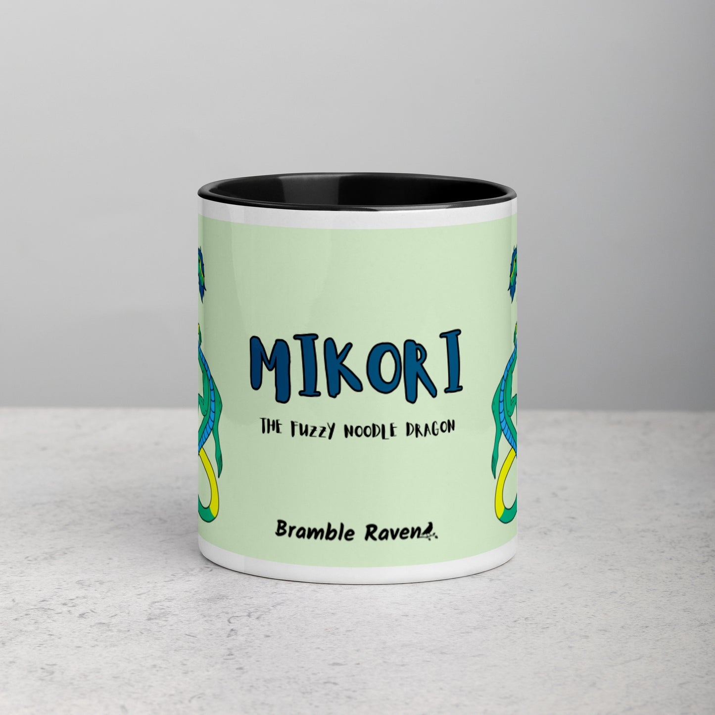 11 ounce white ceramic mug. Black inside and handle. Features double-sided image of Mikori the Fuzzy Noodle Dragon. Shown on tabletop. Front view shows Mikori the Fuzzy Noodle Dragon text and Bramble Raven logo.