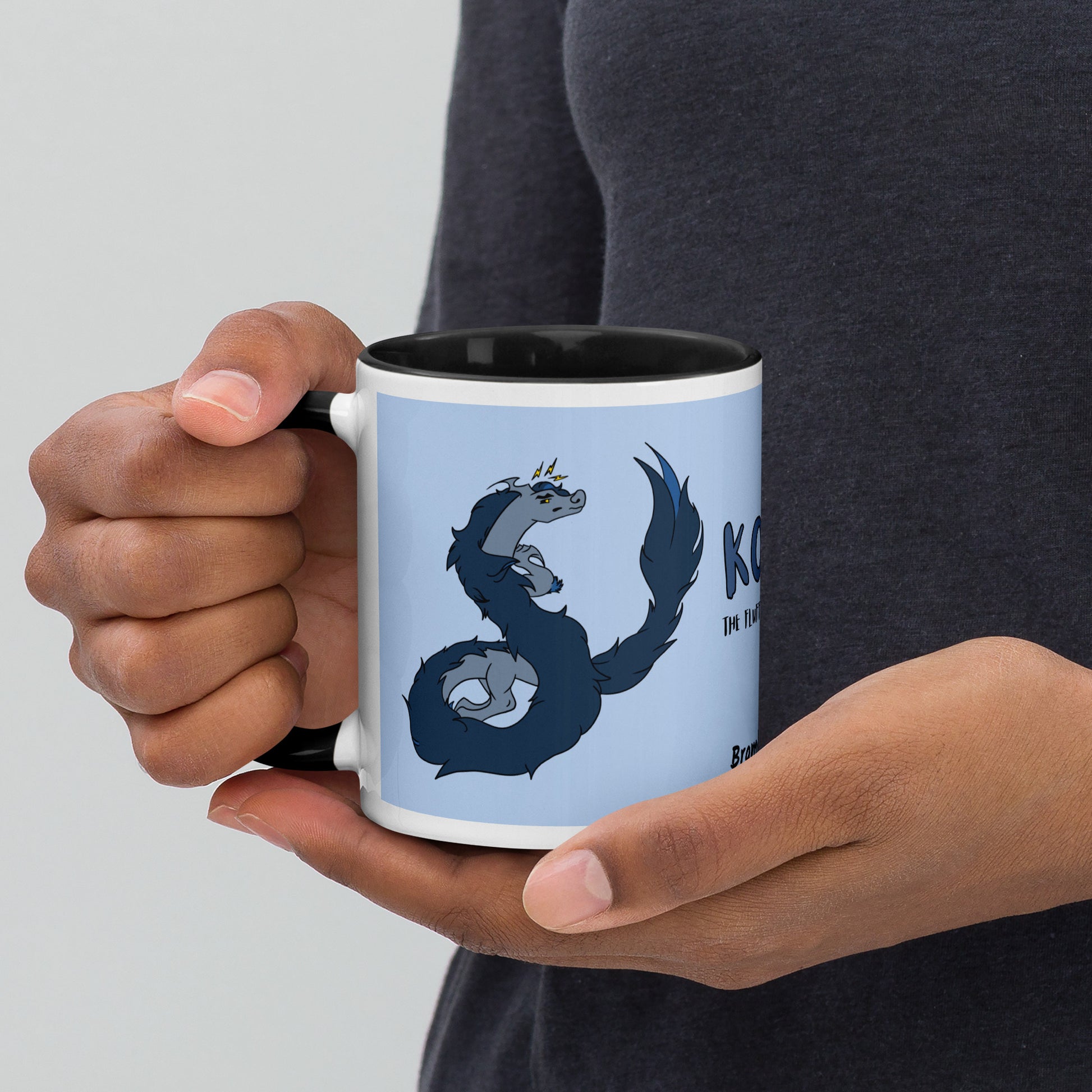 11 ounce ceramic mug featuring a double-sided image of angry Korvo the Fuzzy Noodle dragon against a light blue background.  Black inside and handle. Sitting in hands of model.