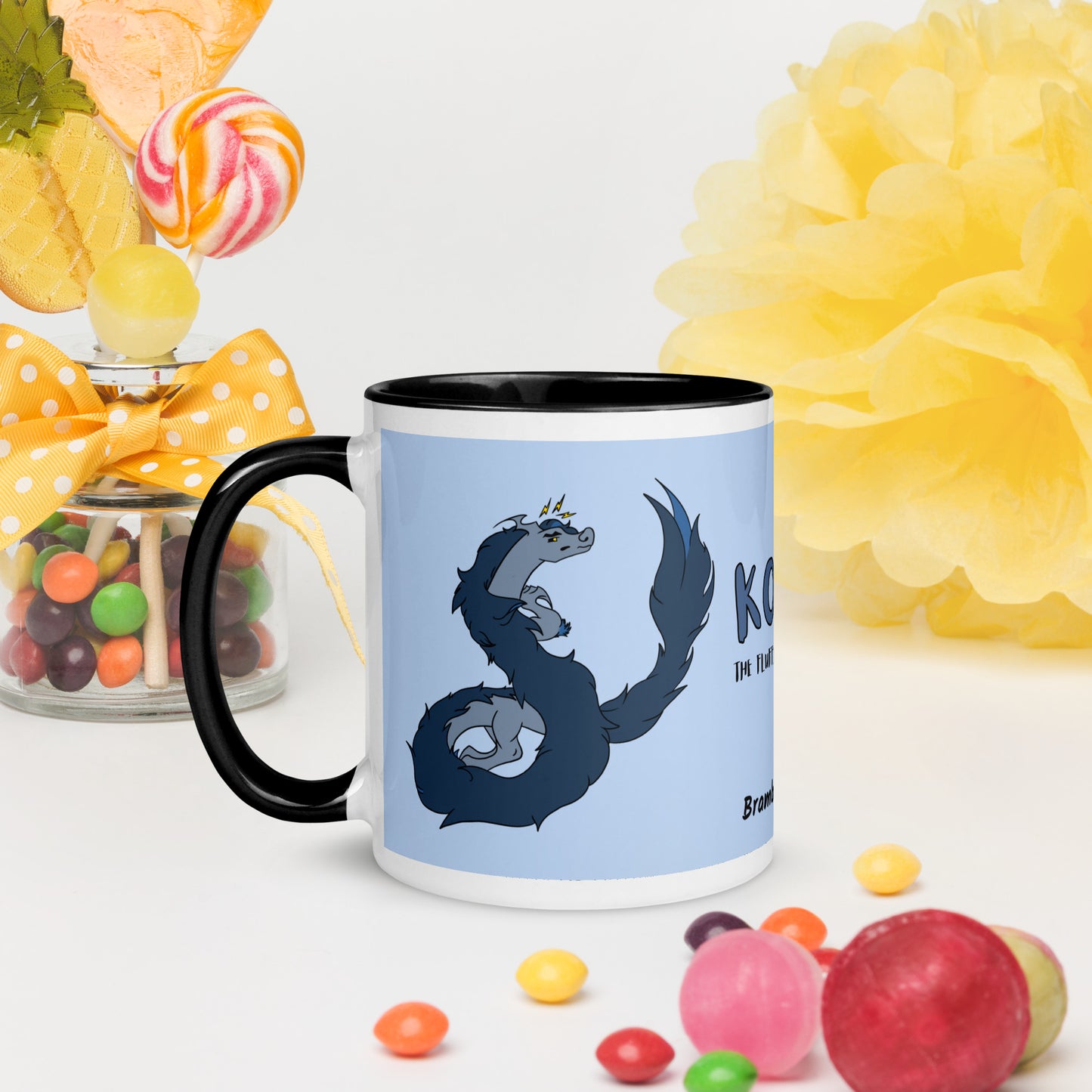 11 ounce ceramic mug featuring a double-sided image of angry Korvo the Fuzzy Noodle dragon against a light blue background.  Black inside and handle. Sitting on tabletop surrounded by candy and decorations.