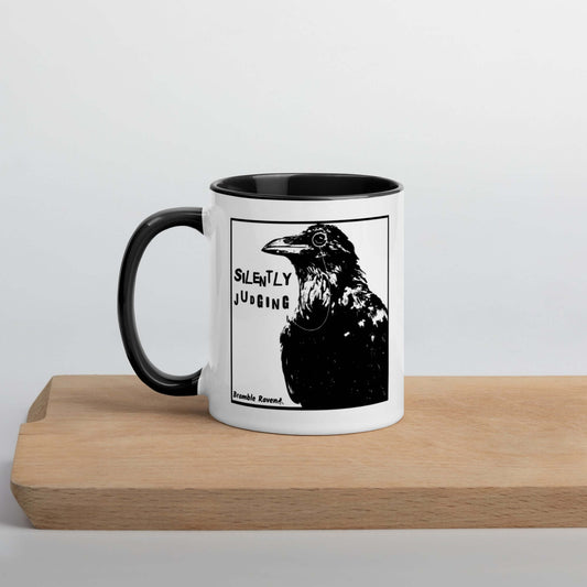 White ceramic mug with silently judging text by black crow wearing a monocle. Double-sided image on 11 oz. mug. Has black handle and inside. Shown on serving board.