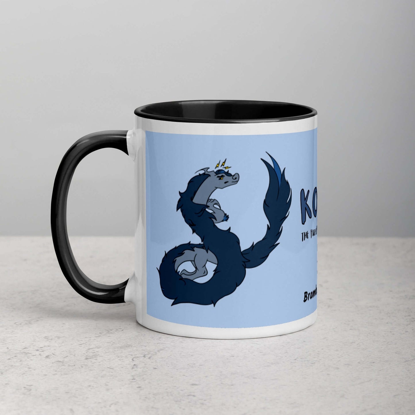 11 ounce ceramic mug featuring a double-sided image of angry Korvo the Fuzzy Noodle dragon against a light blue background.  Black inside and handle. Sitting on tabletop.