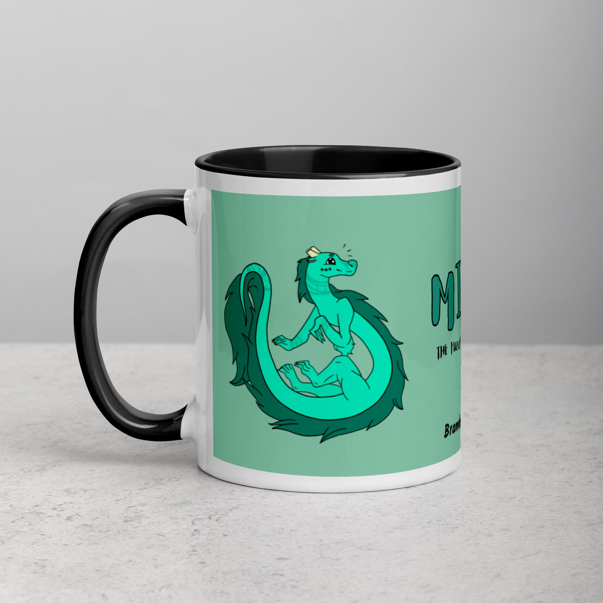 11 ounce white ceramic mug. Black inside and handle. Features double-sided image of Minty the Fuzzy Noodle Dragon on a green background. Shown on tabletop.