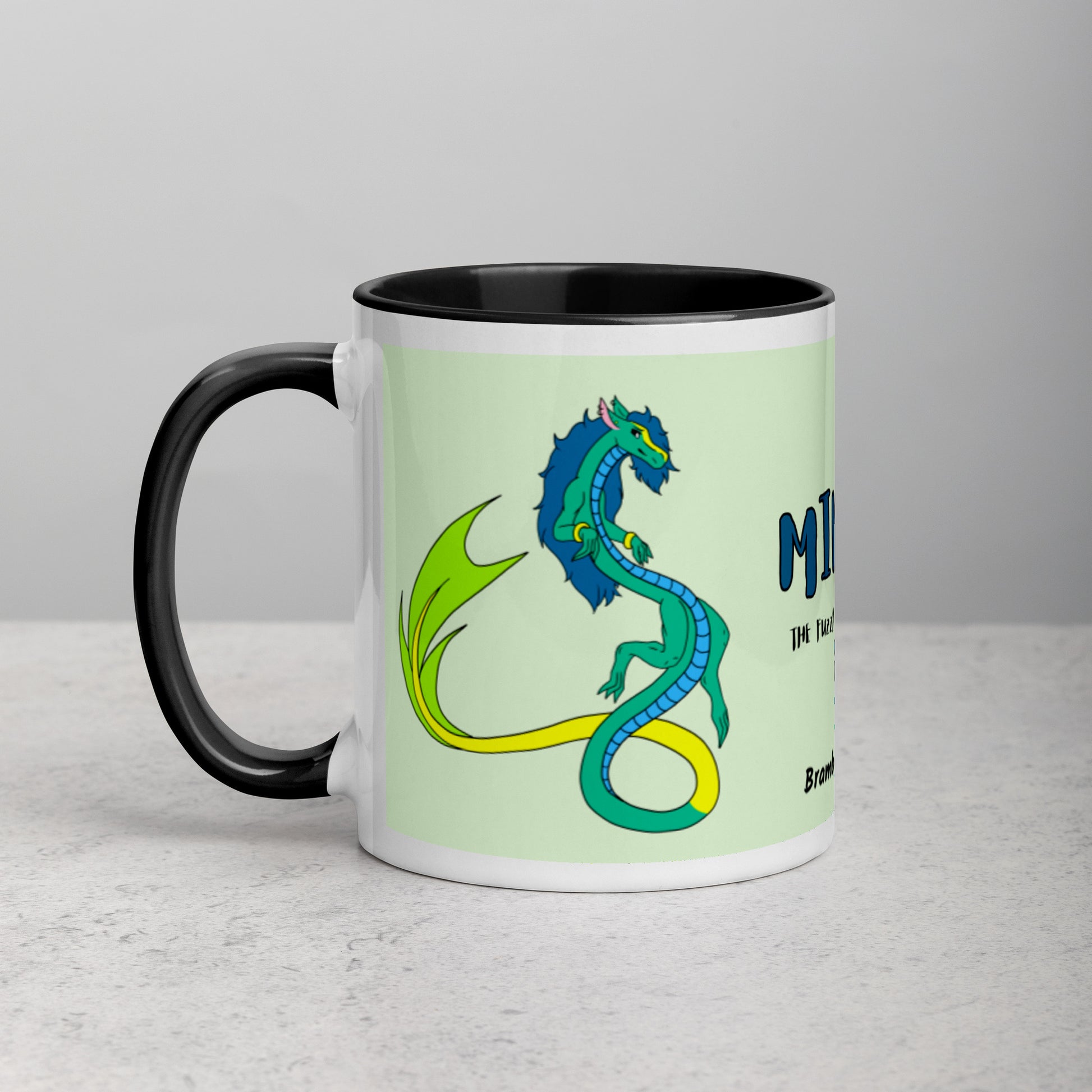 11 ounce white ceramic mug. Black inside and handle. Features double-sided image of Mikori the Fuzzy Noodle Dragon. Shown on tabletop.