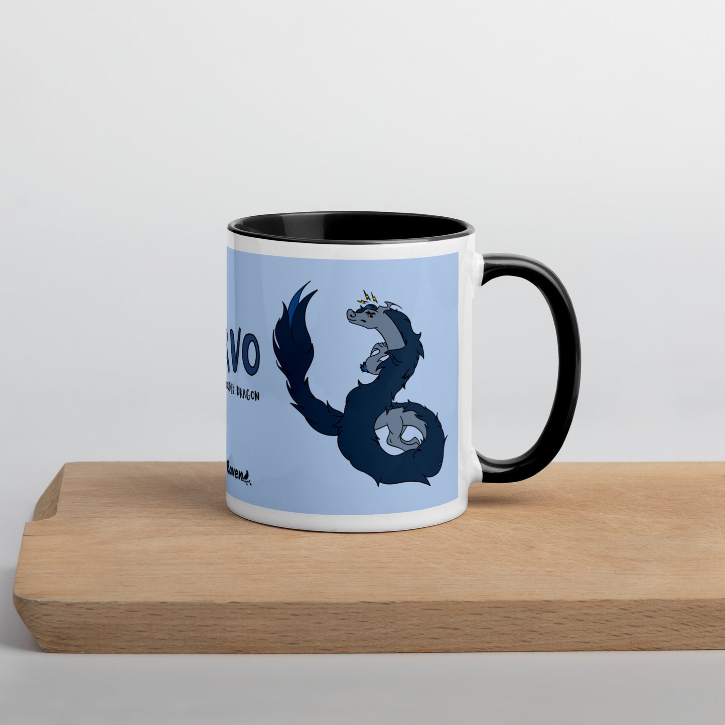 11 ounce ceramic mug featuring a double-sided image of angry Korvo the Fuzzy Noodle dragon against a light blue background.  Black inside and handle. Sitting on serving board with handle facing right.