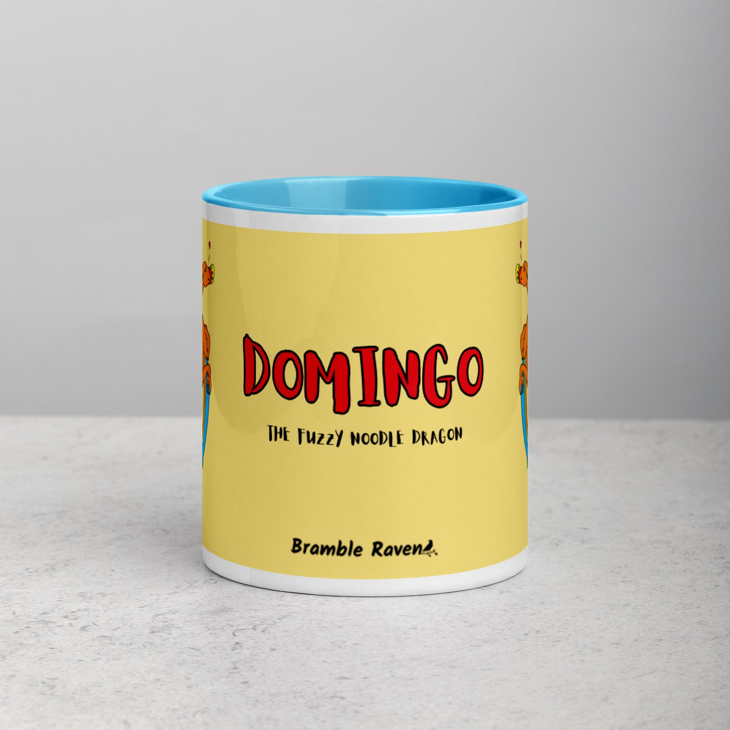 11 ounce white ceramic mug featuring a double-sided image of Domingo the Fuzzy Noodle Dragon against a light yellow background. Blue inside and handle color. Shown on tabletop. Front view shows Domingo the Fuzzy Noodle Dragon text and Bramble Raven logo.
