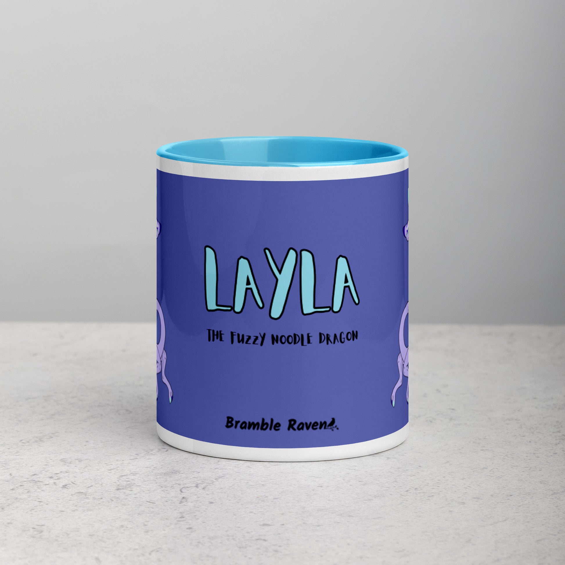 11 ounce white ceramic mug. Blue inside and handle. Features double-sided image of Layla the Lavender Fuzzy Noodle Dragon. Shown on tabletop. Front view shows Layla the Fuzzy Noodle Dragon text and Bramble Raven logo.