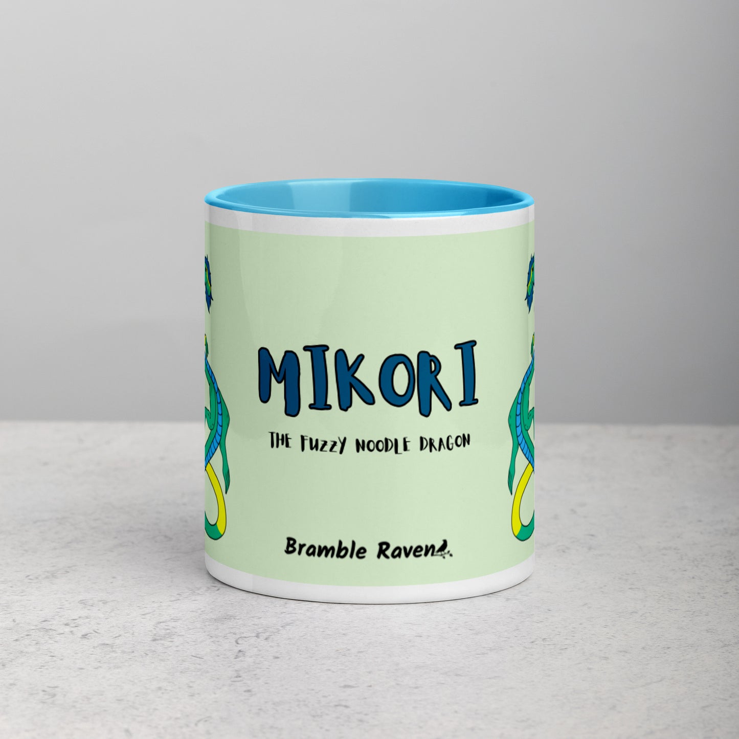 11 ounce white ceramic mug. Blue inside and handle. Features double-sided image of Mikori the Fuzzy Noodle Dragon. Shown on tabletop. Front view shows Mikori the Fuzzy Noodle Dragon text and Bramble Raven logo.