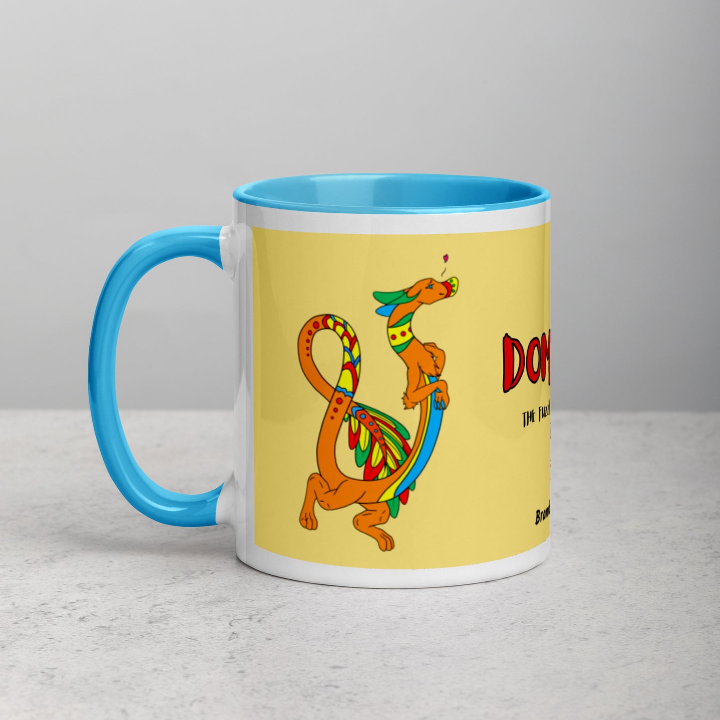 11 ounce white ceramic mug featuring a double-sided image of Domingo the Fuzzy Noodle Dragon against a light yellow background. Blue inside and handle color. Shown on tabletop.