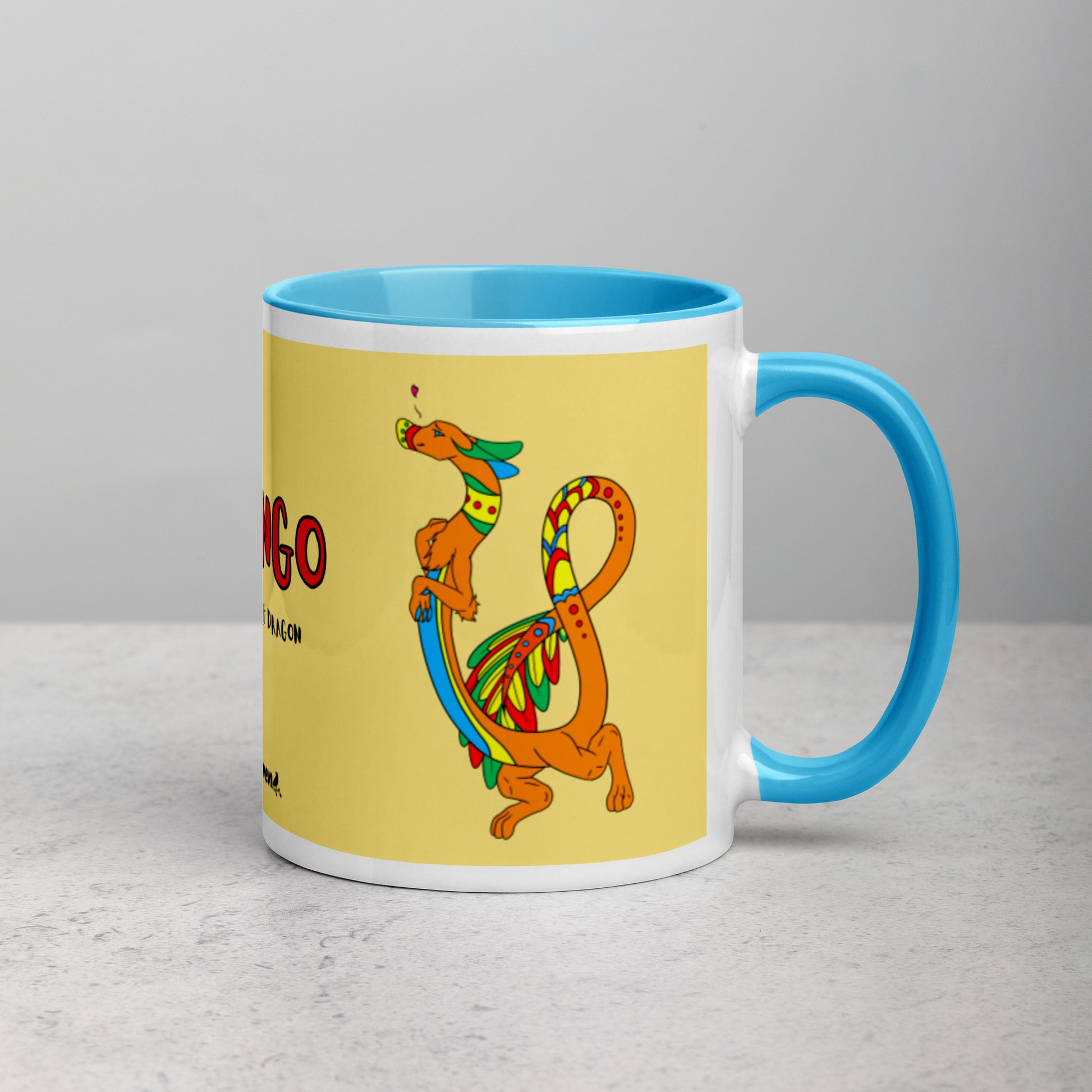 11 ounce white ceramic mug featuring a double-sided image of Domingo the Fuzzy Noodle Dragon against a light yellow background. Blue inside and handle color. Shown on tabletop with handle facing right.
