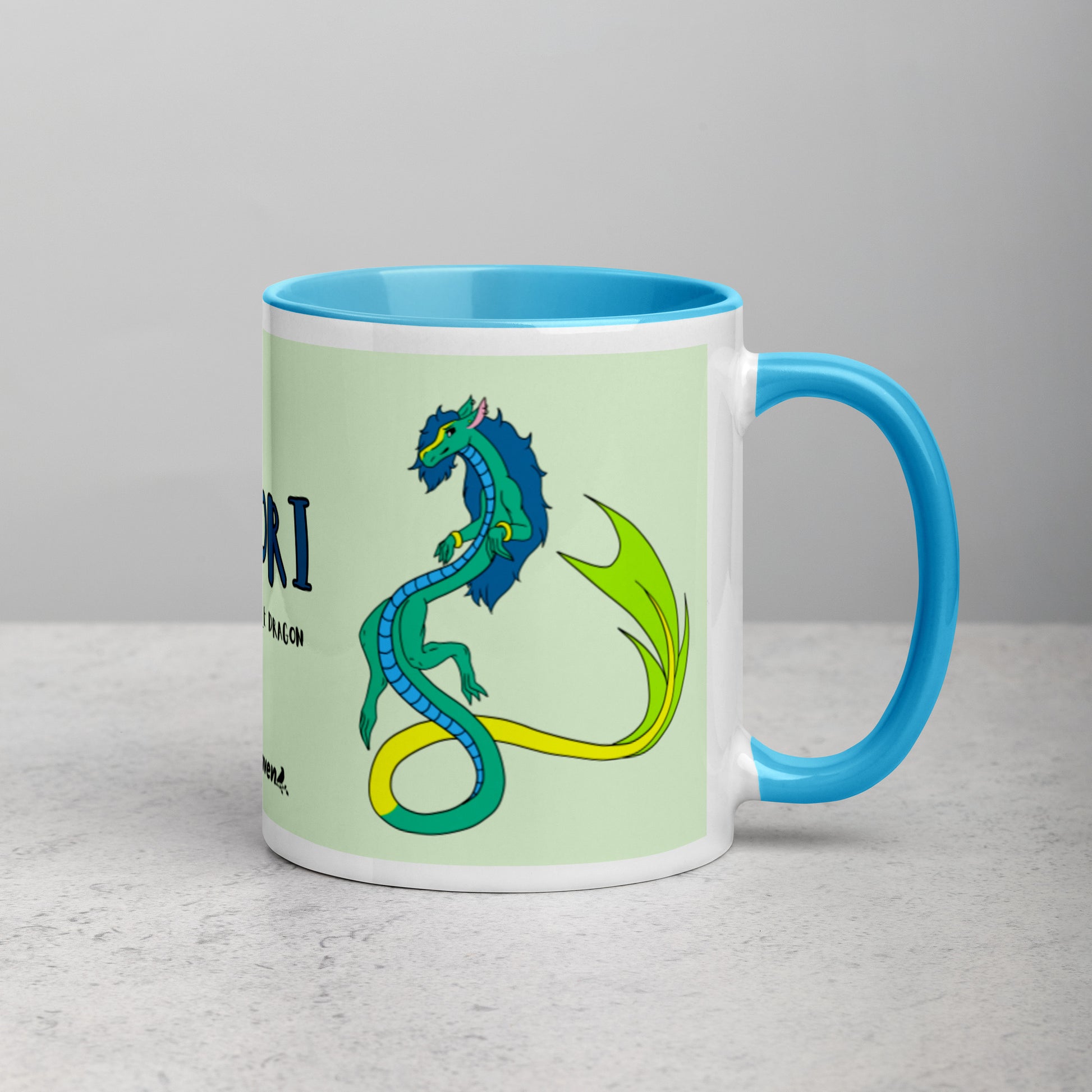 11 ounce white ceramic mug. Blue inside and handle. Features double-sided image of Mikori the Fuzzy Noodle Dragon. Shown on tabletop with handle facing right.