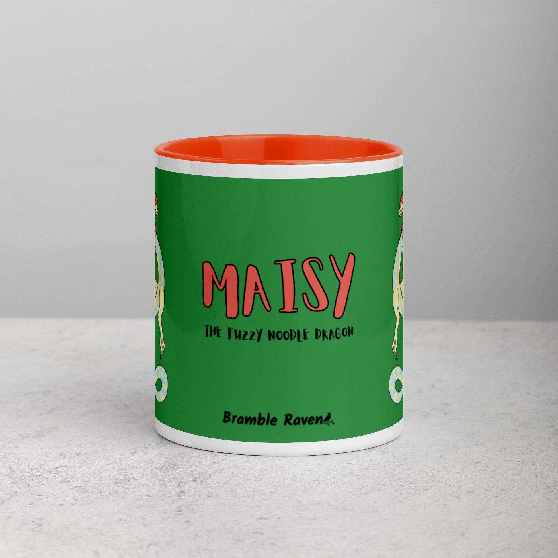 11 ounce white ceramic mug. Orange inside and handle. Features double-sided image of Maisy the Fuzzy Noodle Dragon with a slice of pizza. Shown on tabletop. Front view of mug shows Maisy the Fuzzy Noodle Dragon text and Bramble Raven logo.