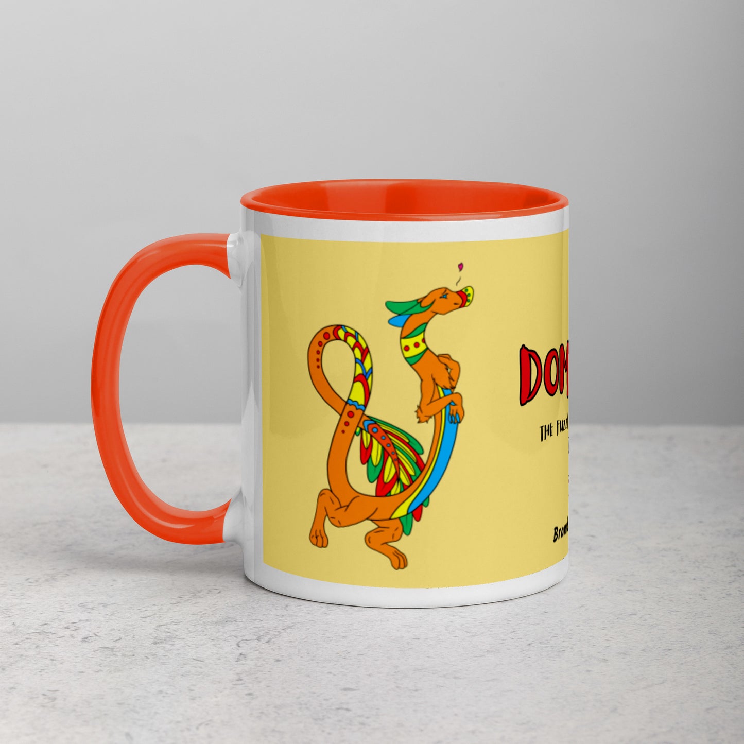 11 ounce white ceramic mug featuring a double-sided image of Domingo the Fuzzy Noodle Dragon against a light yellow background. Orange inside and handle color. Shown on tabletop.