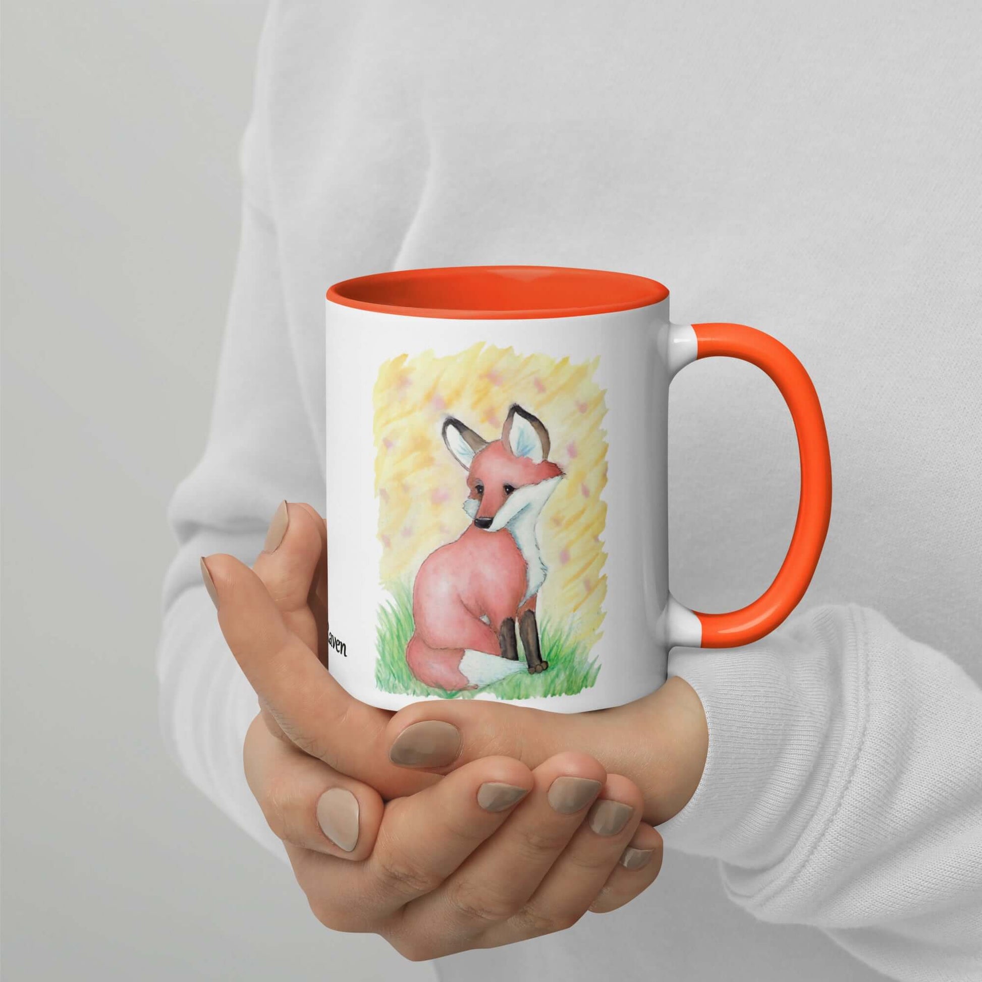 11 oz white ceramic mug with orange inside and handle. Features double-sided image of original watercolor painting of a fox in the grass against a yellow backdrop. Shown in model's hands.