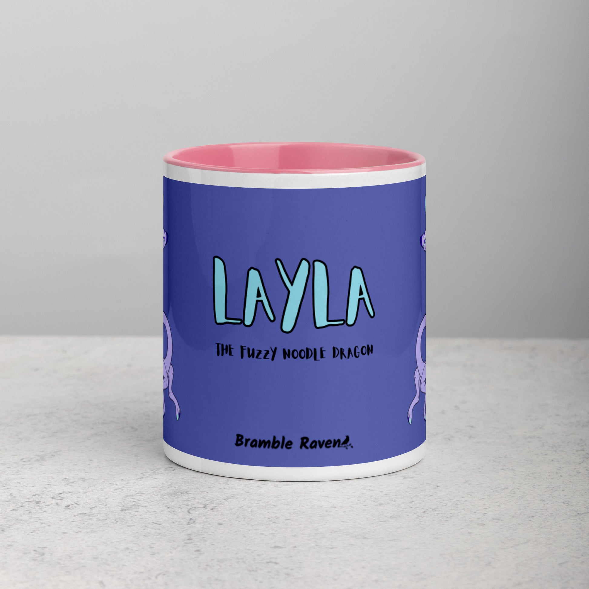 11 ounce white ceramic mug. Pink inside and handle. Features double-sided image of Layla the Lavender Fuzzy Noodle Dragon. Shown on tabletop. Front view shows Layla the Fuzzy Noodle Dragon text and Bramble Raven logo.