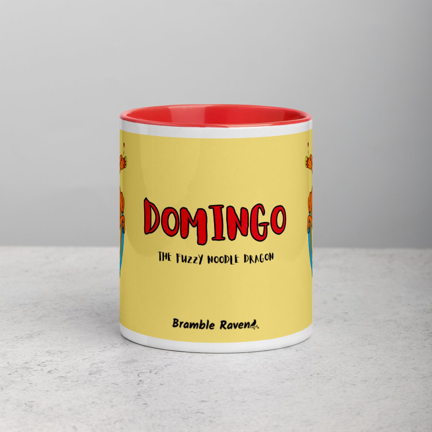 11 ounce white ceramic mug featuring a double-sided image of Domingo the Fuzzy Noodle Dragon against a light yellow background. Red inside and handle color. Shown on tabletop. Front view shows Domingo the Fuzzy Noodle Dragon text and Bramble Raven logo.