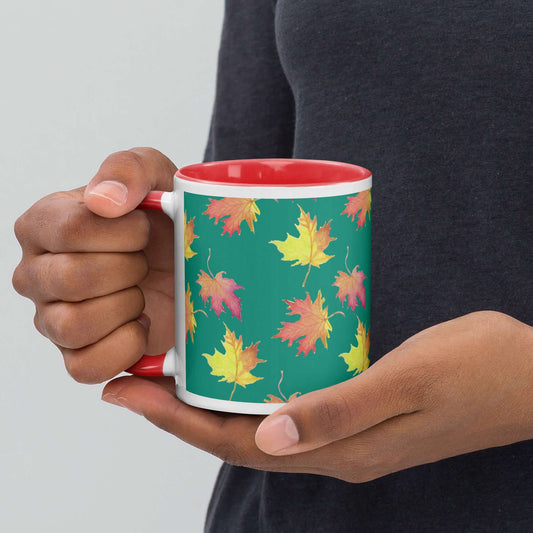 White ceramic mug with red color inside and on handle. Features pattern of watercolor fall leaves on a green background. Holds 11 ounces. Shown in the hands of a model.