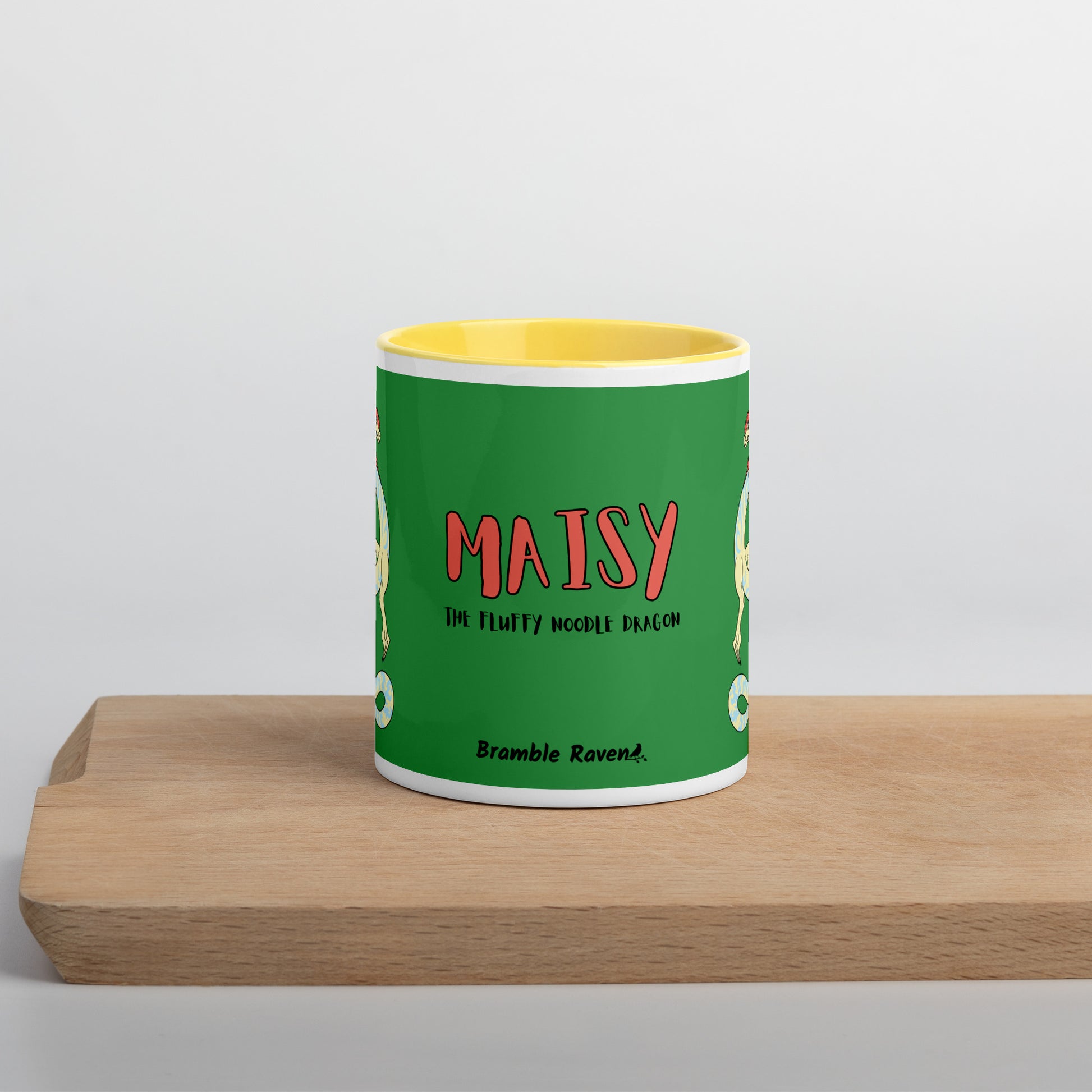 11 ounce white ceramic mug. Yellow inside and handle. Features double-sided image of Maisy the Fuzzy Noodle Dragon with a slice of pizza.  Front view shows Maisy the Fluffy Noodle Dragon text with Bramble Raven logo.