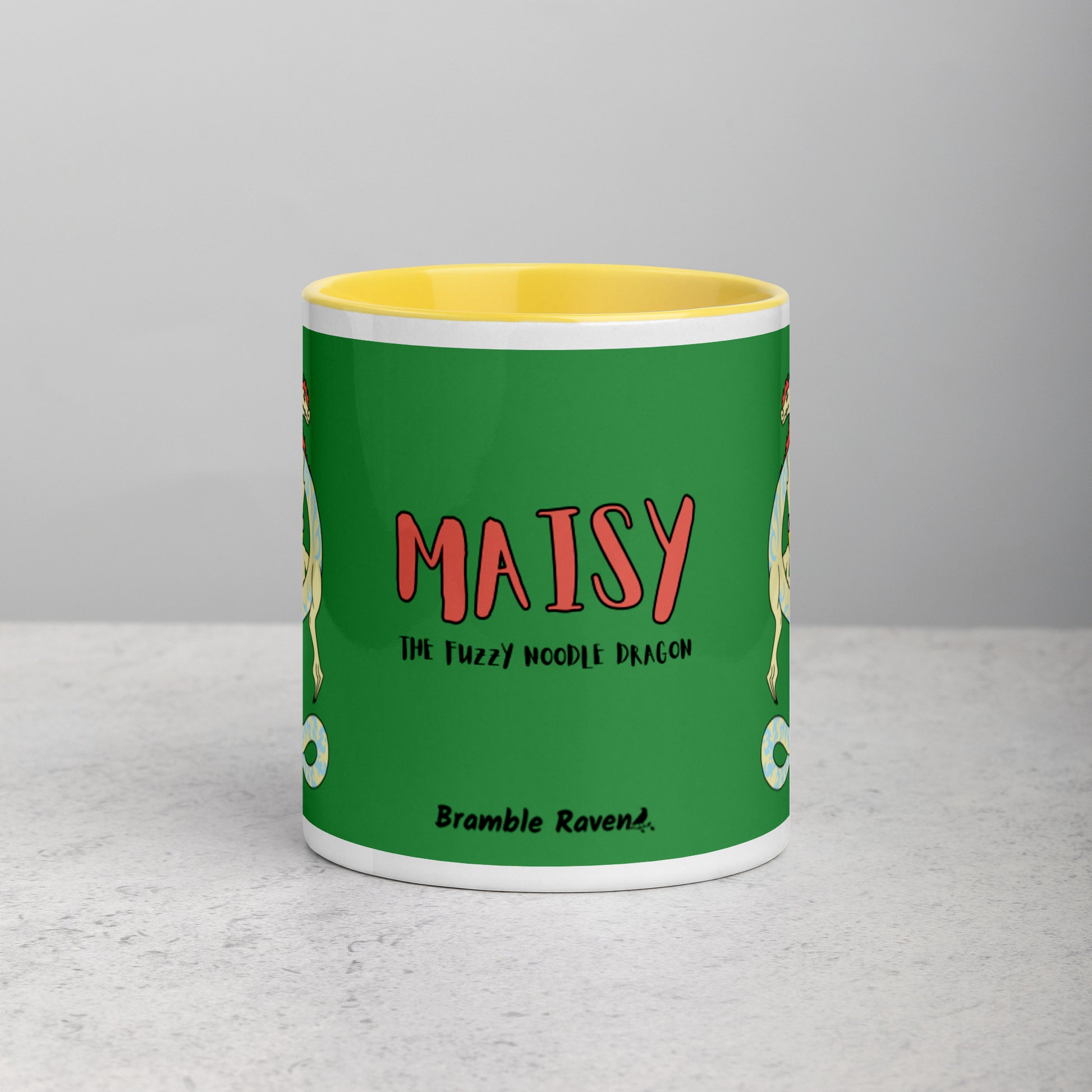 11 ounce white ceramic mug. Yellow inside and handle. Features double-sided image of Maisy the Fuzzy Noodle Dragon with a slice of pizza. Shown on tabletop. Front view of mug shows Maisy the Fuzzy Noodle Dragon text and Bramble Raven logo.