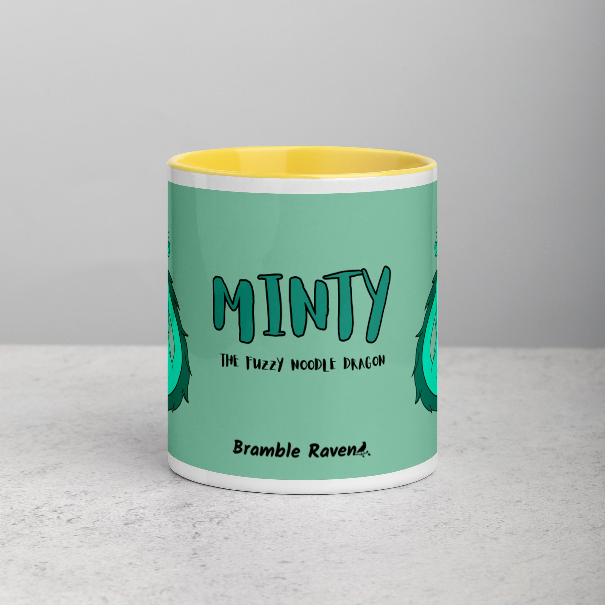 11 ounce white ceramic mug. Yellow inside and handle. Features double-sided image of Minty the Fuzzy Noodle Dragon on a green background. Shown on tabletop. Front view shows Minty the Fuzzy Noodle Dragon text and Bramble Raven logo.
