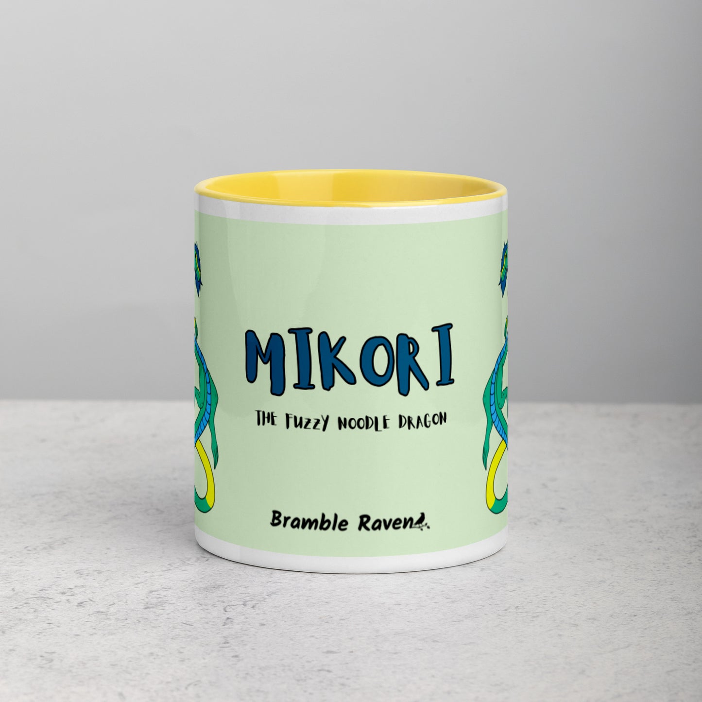 11 ounce white ceramic mug. Yellow inside and handle. Features double-sided image of Mikori the Fuzzy Noodle Dragon. Shown on tabletop. Front view shows Mikori the Fuzzy Noodle Dragon text and Bramble Raven logo.