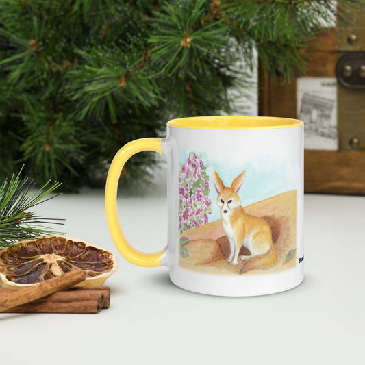 White ceramic 11 ounce mug with yellow handle and inside. Features original watercolor painting of a fennec fox in the desert on both sides. Shown next to pine boughs and dried citrus slices and cinnamon sticks.
