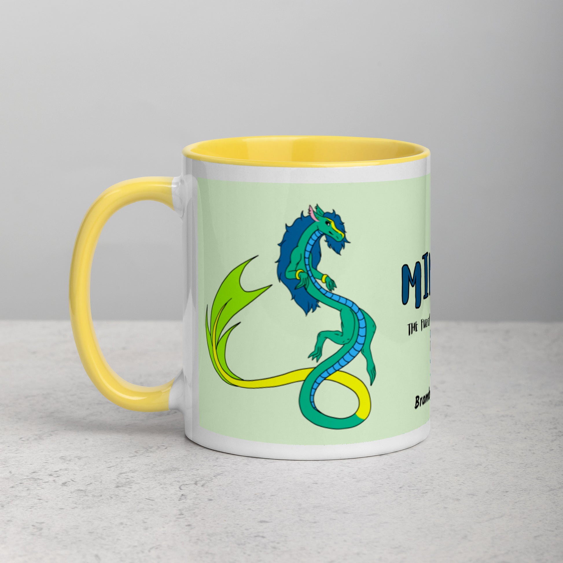 11 ounce white ceramic mug. Yellow inside and handle. Features double-sided image of Mikori the Fuzzy Noodle Dragon. Shown on tabletop.