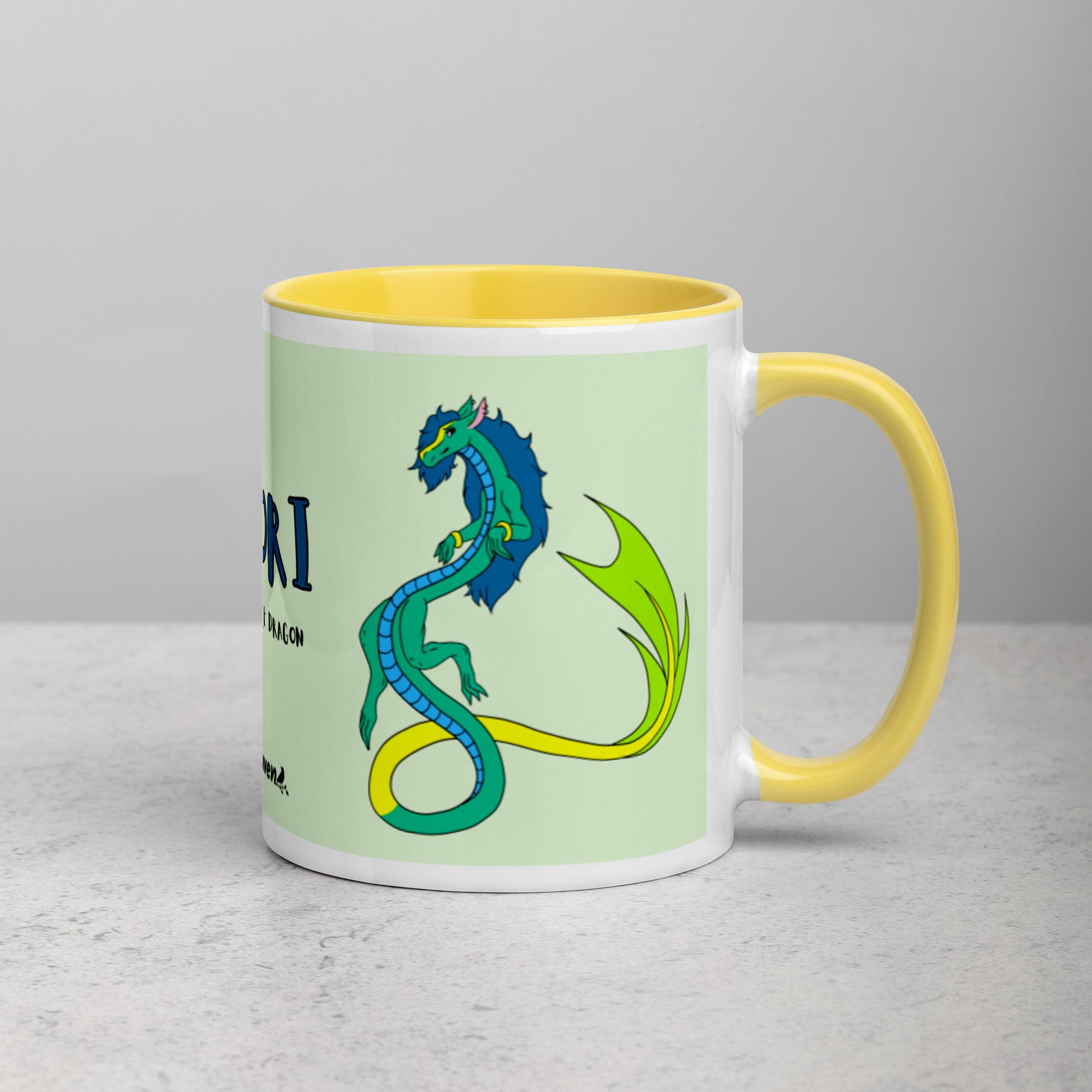 11 ounce white ceramic mug. Yellow inside and handle. Features double-sided image of Mikori the Fuzzy Noodle Dragon. Shown on tabletop with handle facing right.