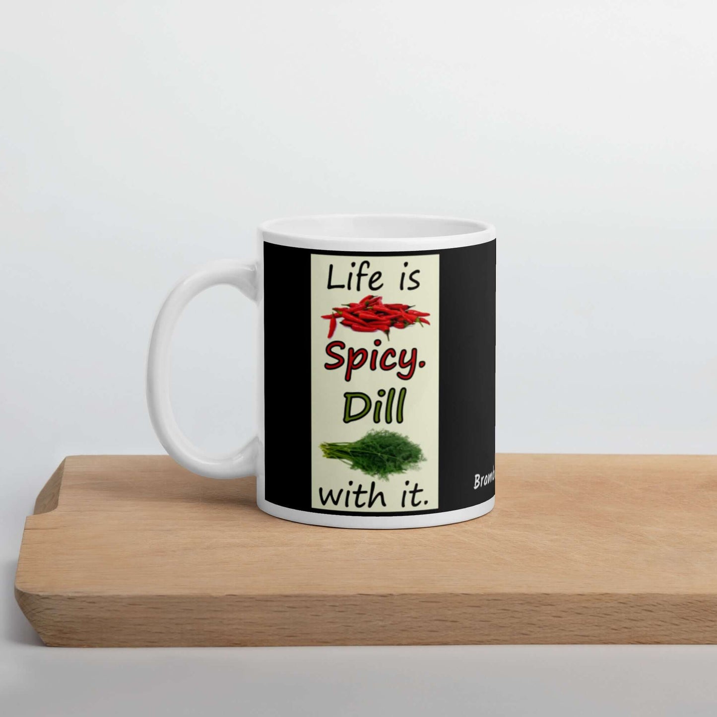 11 ounce white glossy ceramic mug. Life is Spicy. Dill with it text. Image of chili peppers and dill weed on a light yellow rectangle. Black background on mug. Shown on cutting board.