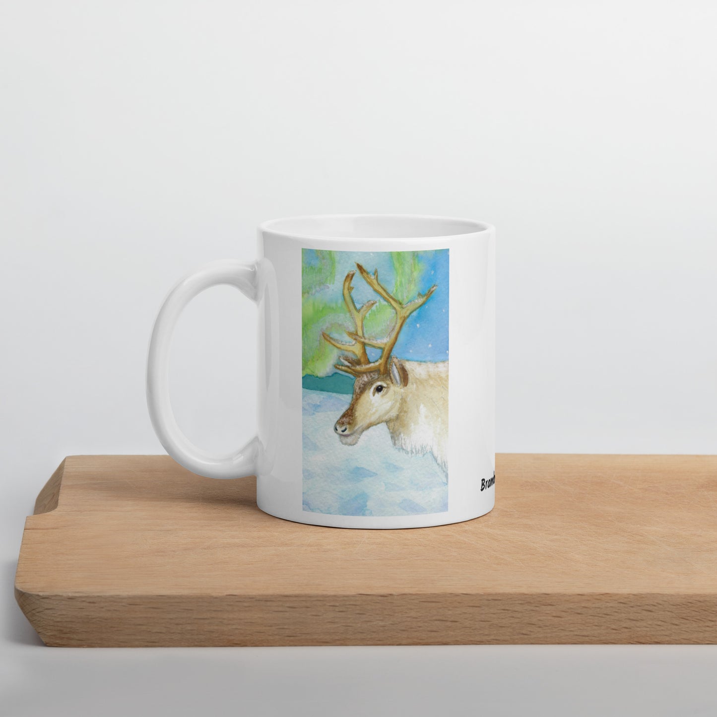 11 ounce ceramic mug featuring Northern Lights reindeer design. Double-sided print. Dishwasher and microwave safe. Shown on wooden cutting board.