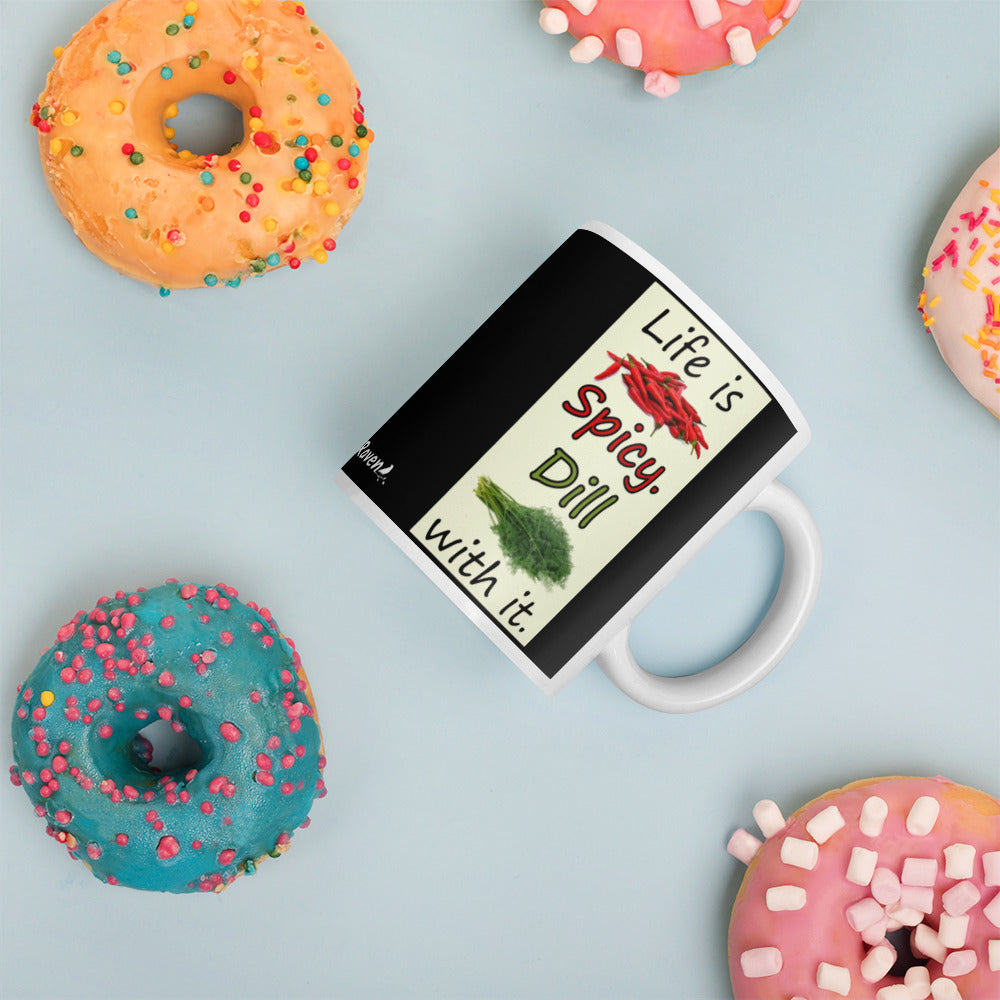 11 ounce white glossy ceramic mug. Life is Spicy. Dill with it text. Image of chili peppers and dill weed on a light yellow rectangle. Black background on mug. Shown laying on tabletop surrounded by donuts.