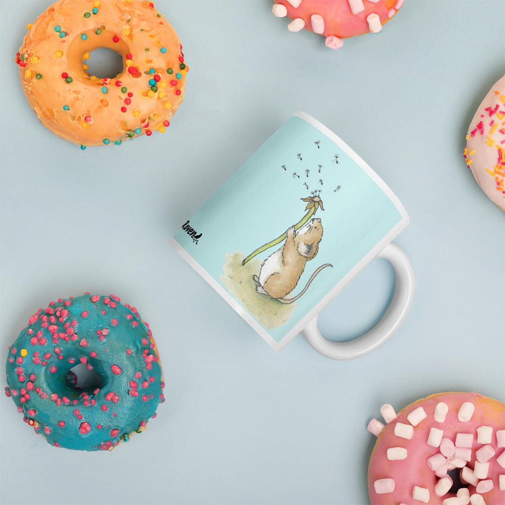 Two-sided image of original Dandelion wish design of cute watercolor mouse blowing dandelion seeds  featured on 11 ounce mug with light blue background. Laying on table surrounded by donuts.