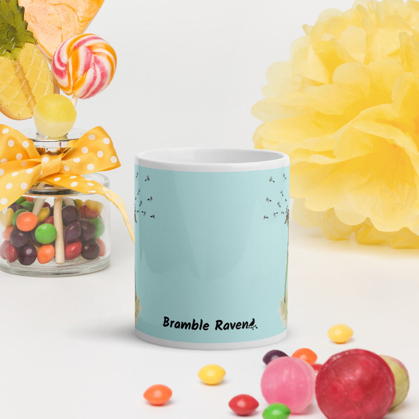 Two-sided image of original Dandelion wish design of cute watercolor mouse blowing dandelion seeds  featured on 11 ounce mug with light blue background. Front view with logo. On table with candy and decorations.