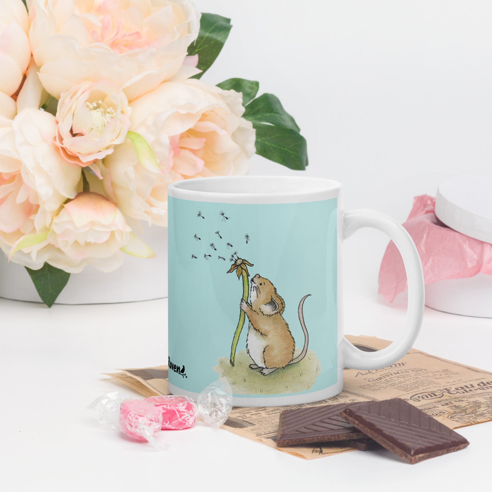 Two-sided image of original Dandelion wish design of cute watercolor mouse blowing dandelion seeds  featured on 11 ounce mug with light blue background. On table with flowers and chocolate.
