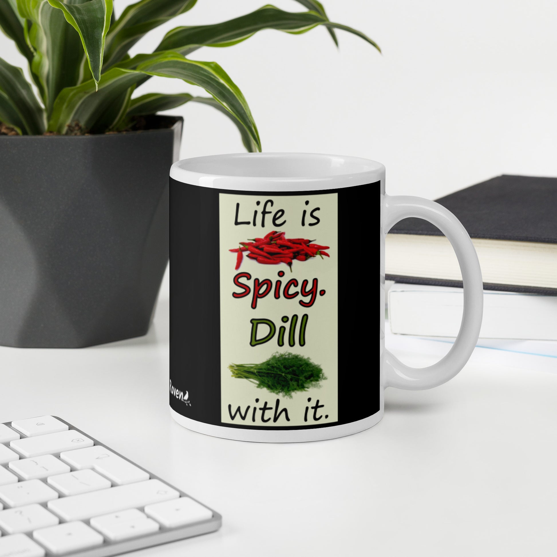 11 ounce white glossy ceramic mug. Life is Spicy. Dill with it text. Image of chili peppers and dill weed on a light yellow rectangle. Black background on mug. Shown on desktop by potted plant and keyboard.