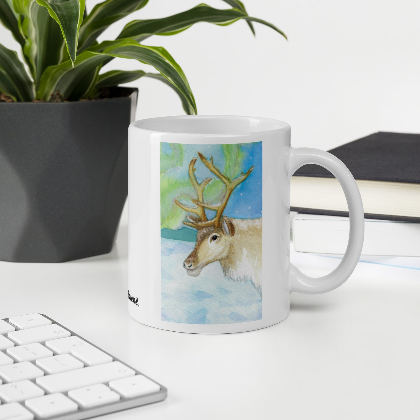 11 ounce ceramic mug featuring Northern Lights reindeer design. Double-sided print. Dishwasher and microwave safe. Shown on a desk by a keyboard, books, and potted plant.