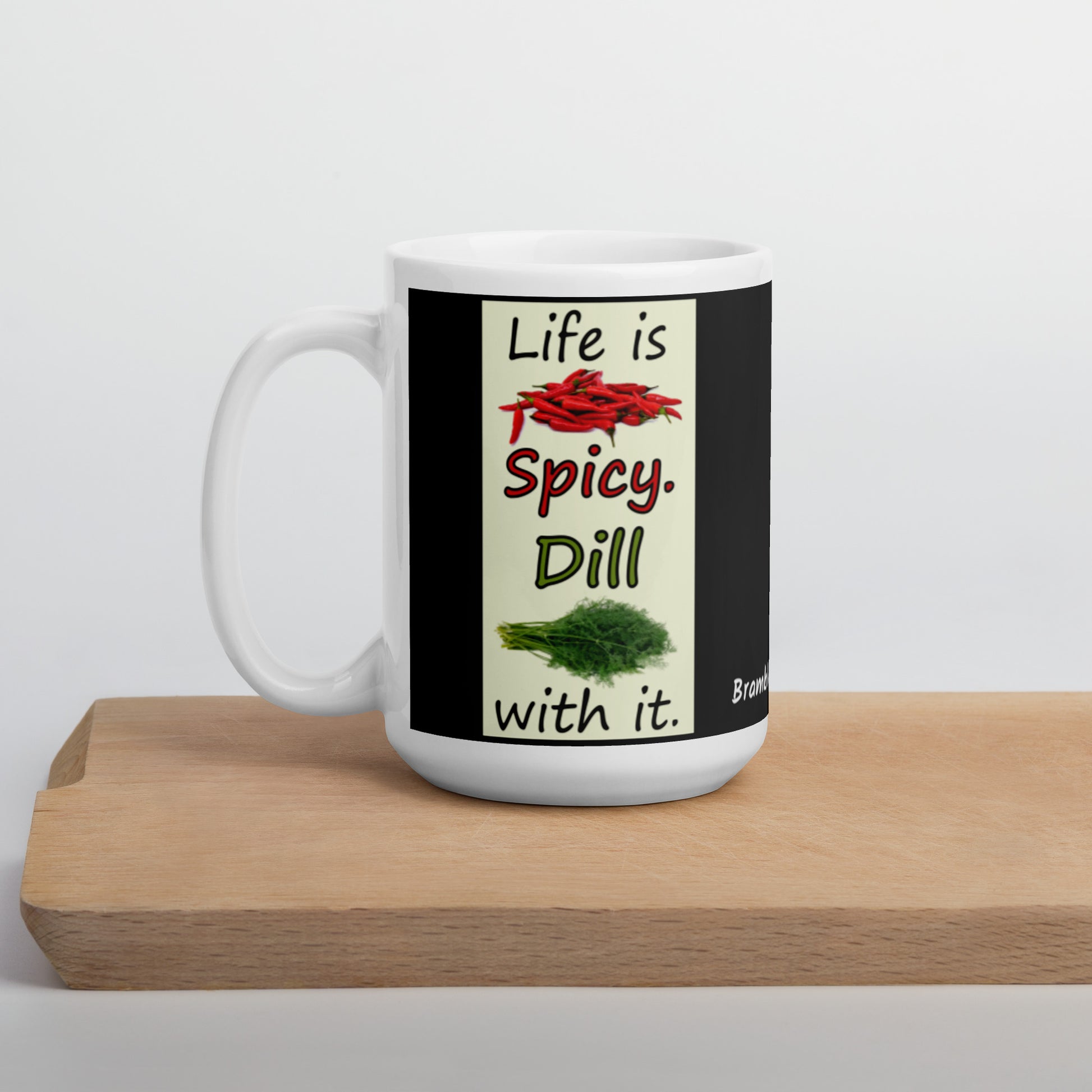 15 ounce white glossy ceramic mug. Life is Spicy. Dill with it text. Image of chili peppers and dill weed on a light yellow rectangle. Black background on mug. Shown on cutting board.