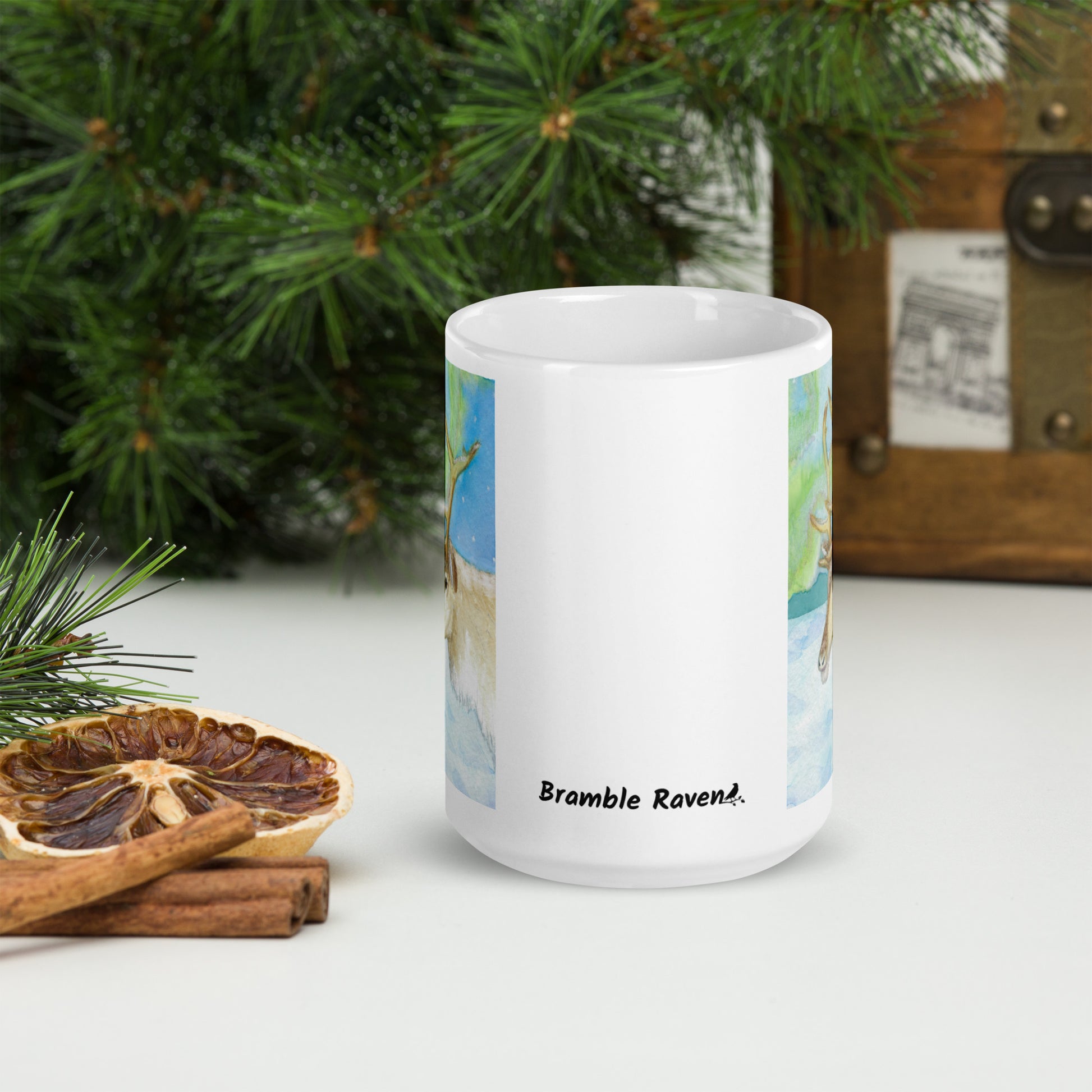 15 ounce ceramic mug featuring Northern Lights reindeer design. Double-sided print. Dishwasher and microwave safe. Front view shows Bramble Raven logo. Shown by dried citrus slices and pine boughs.